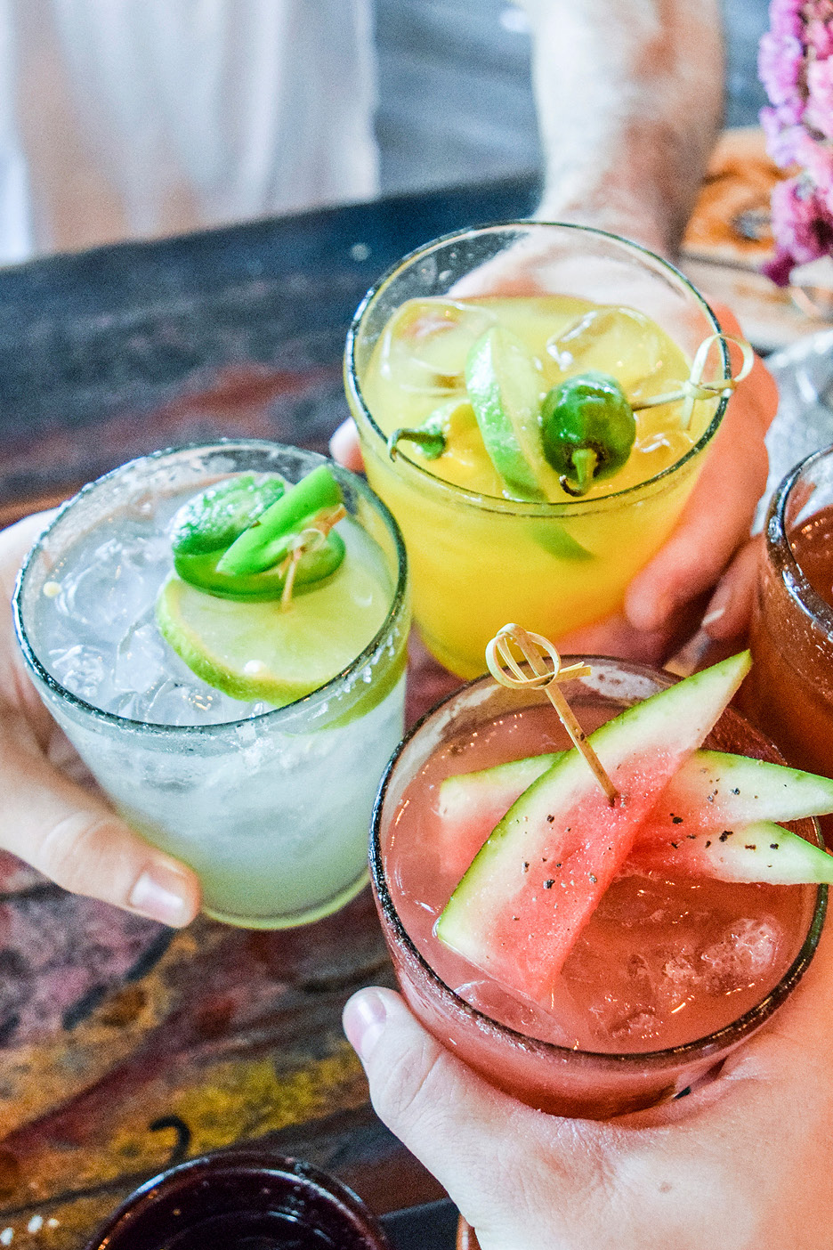 Tre cocktail colorati con guarnizioni di lime, peperoncino e anguria, tenuti in mano.
