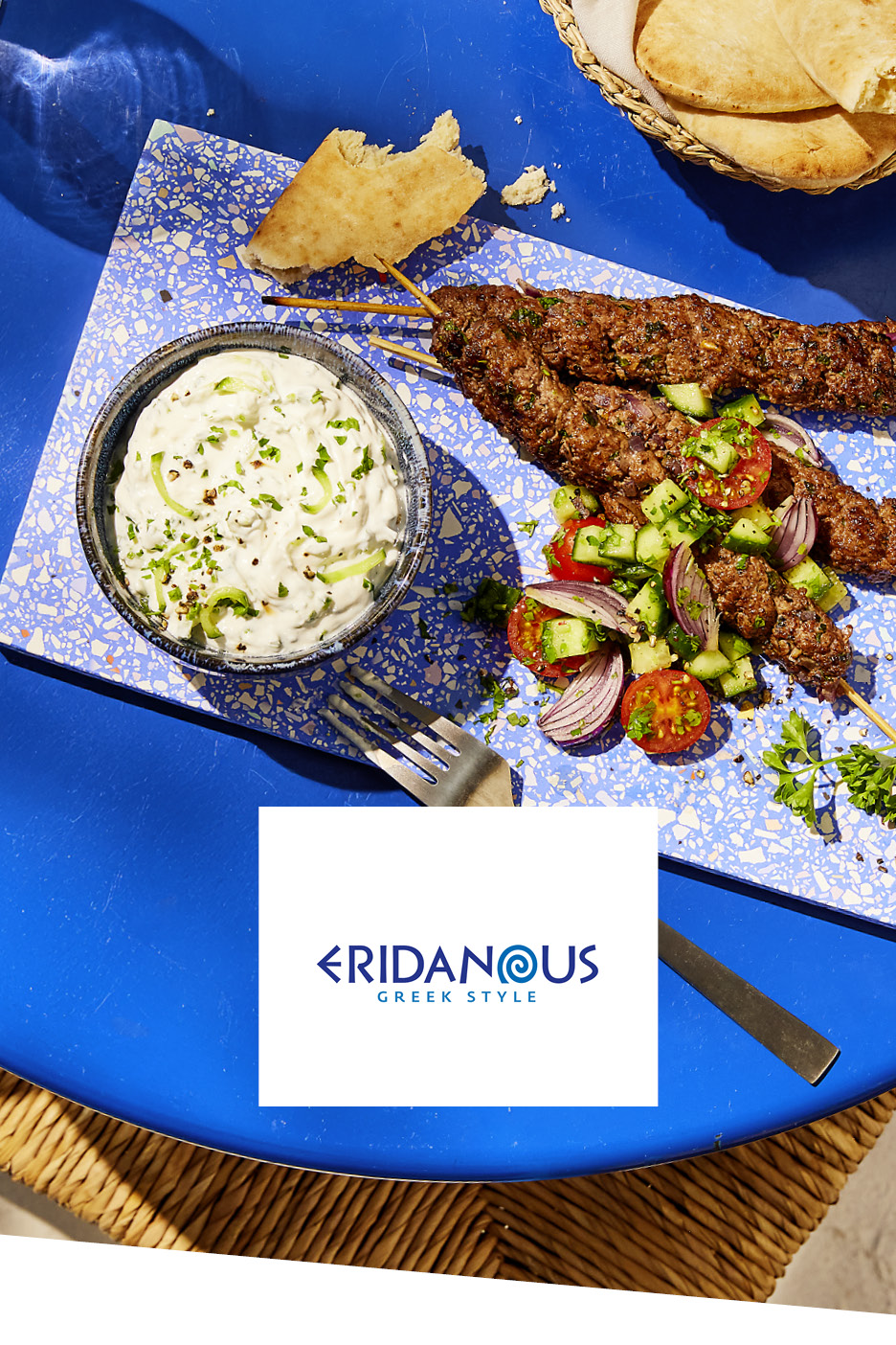 Spiedini di carne con tzatziki, insalata di pomodori e cetrioli, e pane pita su un tavolo blu.
