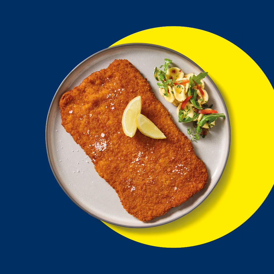 Cotoletta impanata con limone e insalata di pasta su piatto grigio.