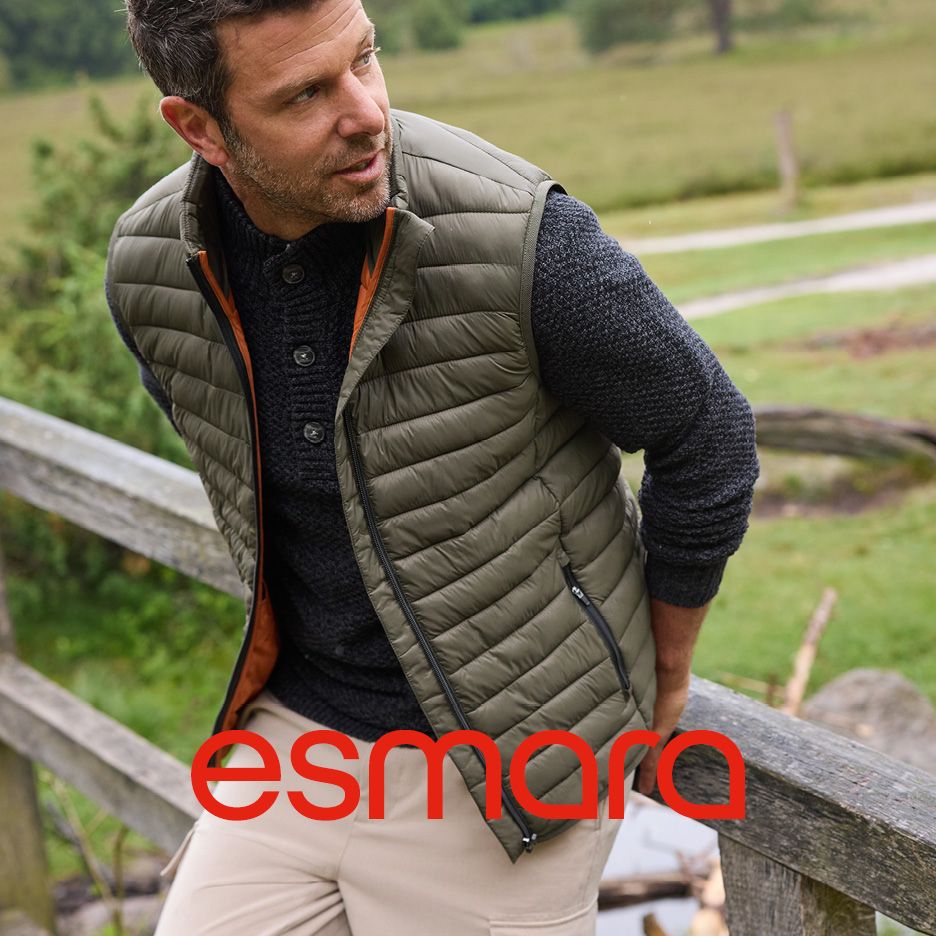 Uomo con gilet trapuntato verde e maglione scuro su un ponte di legno in campagna.