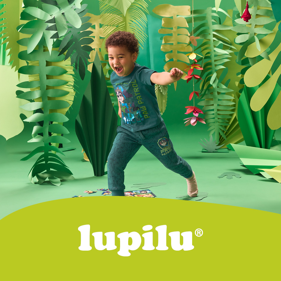 Un bambino sorridente in tuta da gioco verde con stampa di cartoni animati, circondato da piante di carta.
