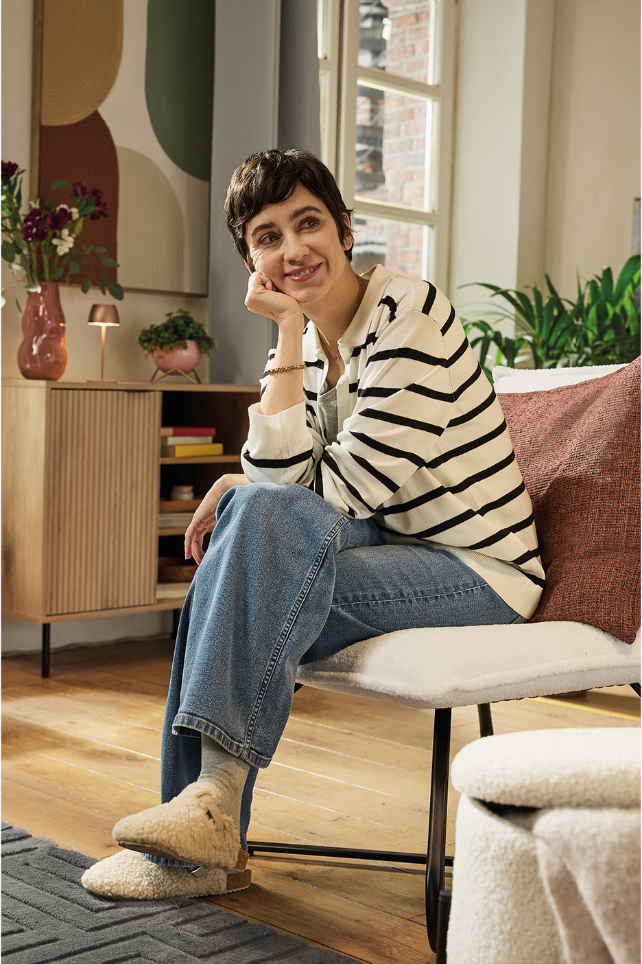 Donna sorridente con maglione a righe e jeans, seduta su una sedia in un soggiorno accogliente.