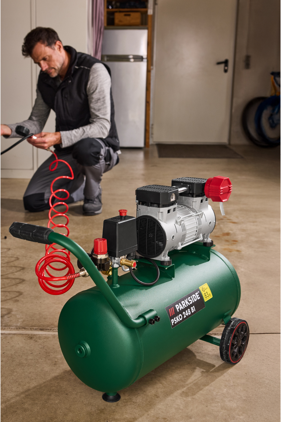 Un uomo si china per collegare un tubo rosso a un compressore d'aria verde con ruote.