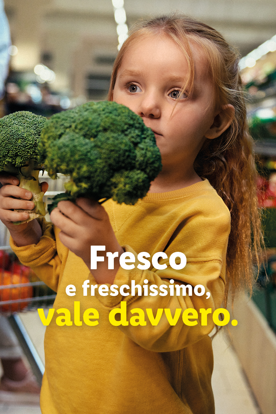 Una bambina con i capelli rossi tiene in mano due broccoli freschi in un supermercato.
