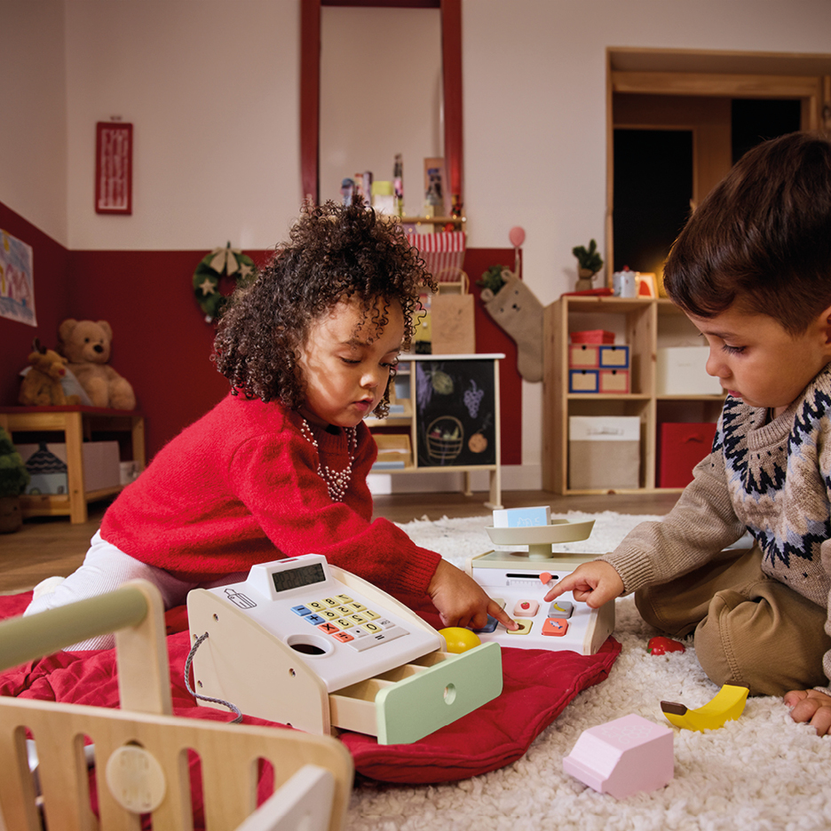 Due bambini giocano con una cassa giocattolo e una bilancia giocattolo su un tappeto.