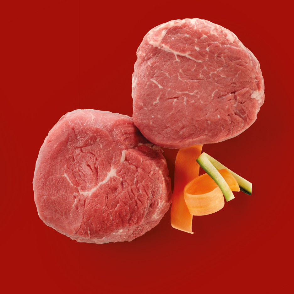 Due bistecche di manzo crude con carote e zucchine su sfondo rosso.