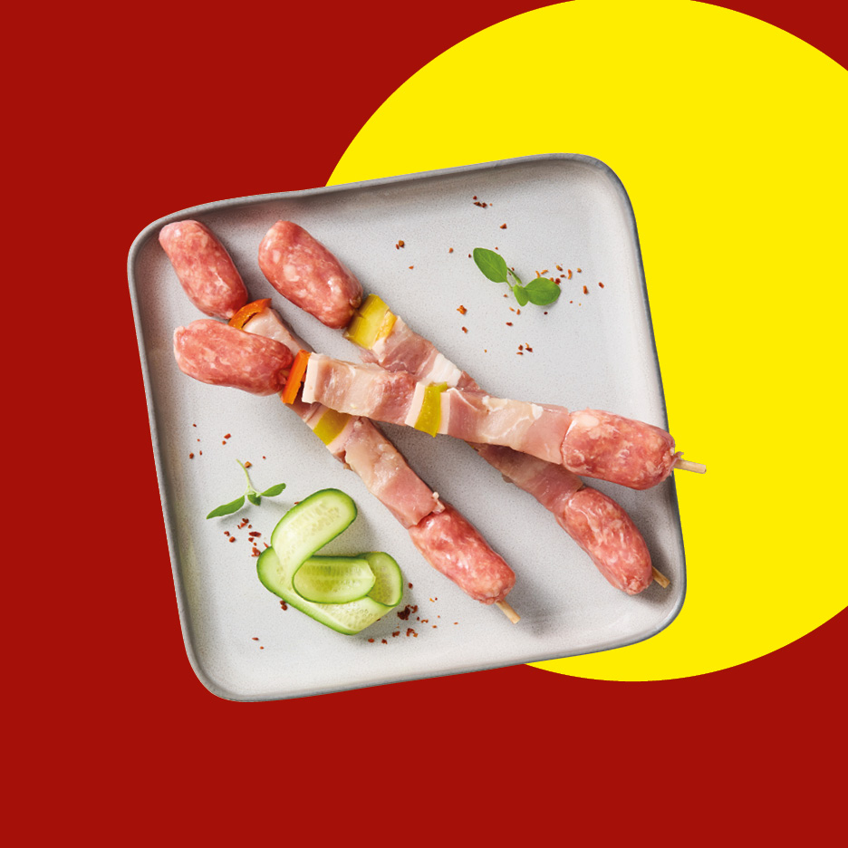 Spiedini di carne cruda con salsiccia e pancetta su un piatto grigio.