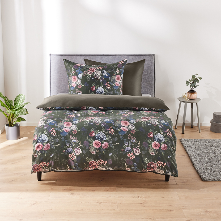 Letto con copripiumino floreale verde scuro, cuscini abbinati e comodino con pianta.