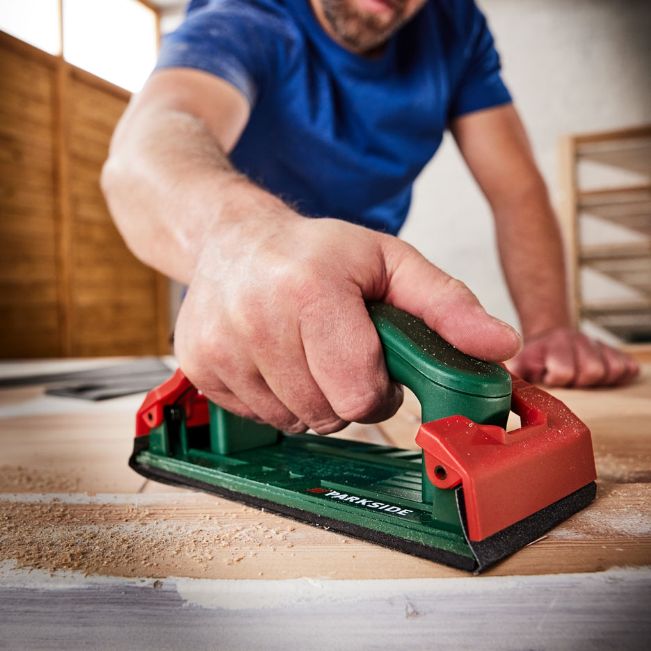 Uomo che leviga una superficie di legno con una levigatrice manuale, con segatura intorno.