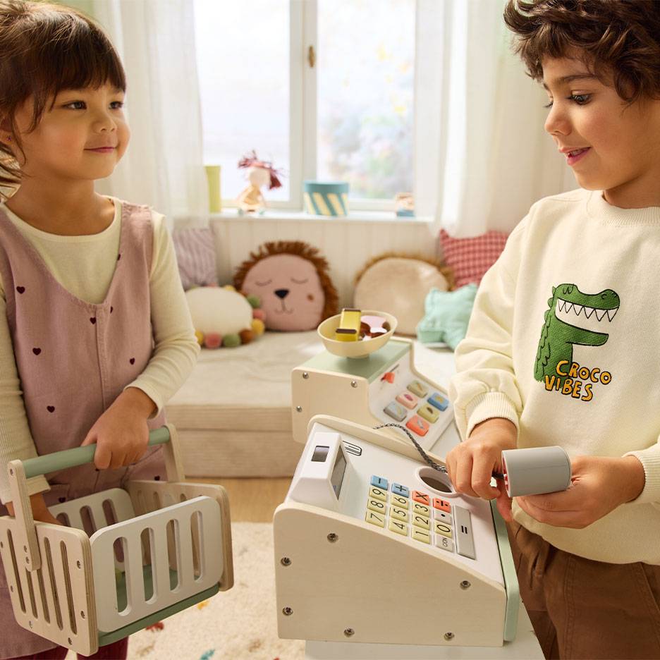 Bambini che giocano al negozio; il bambino usa un registratore di cassa, la bambina porta un cestino.