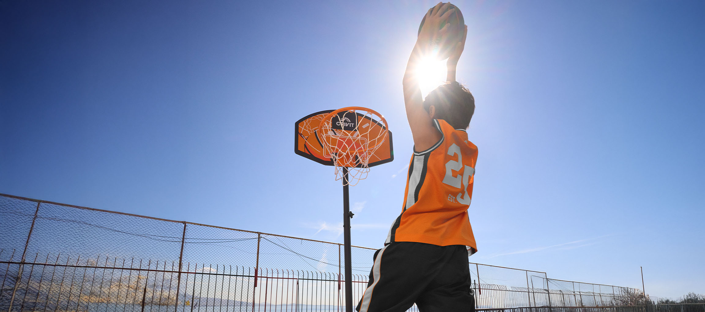Ragazzo con maglia da basket arancione che lancia la palla nel canestro in una giornata di sole.