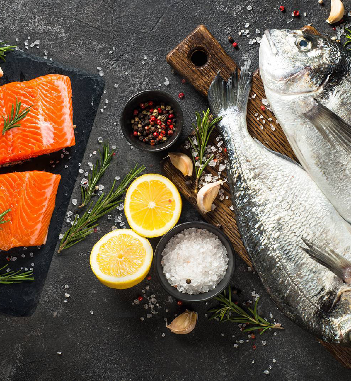 Filetti di salmone e pesce intero fresco con limone, rosmarino, aglio, sale e pepe su sfondo scuro.
