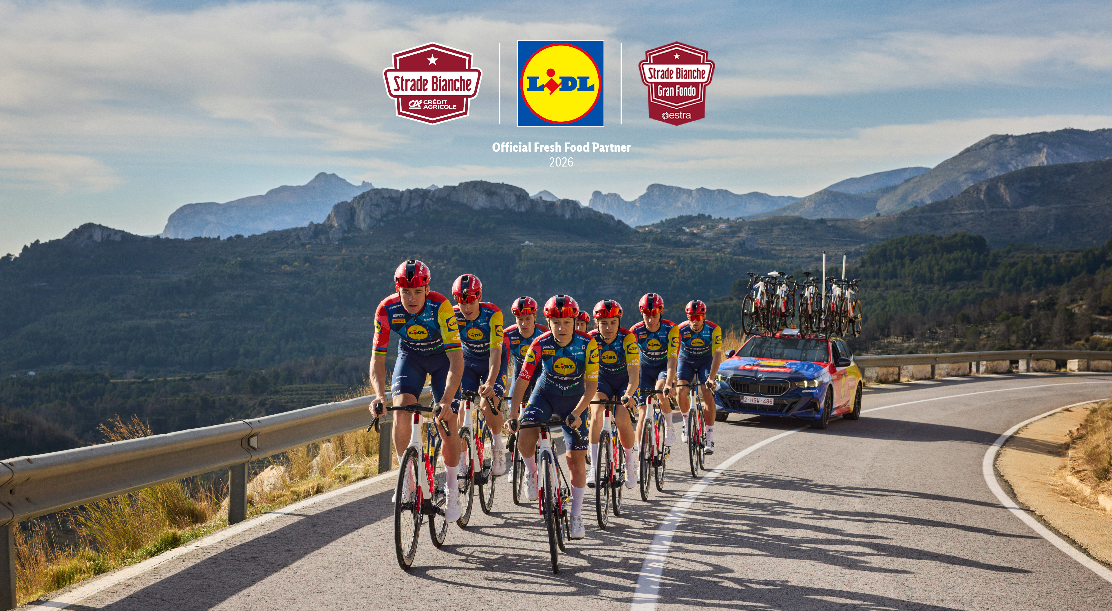 Ciclisti in maglia blu e gialla su una strada di montagna, con un'auto di supporto e loghi di ciclismo e supermercato.