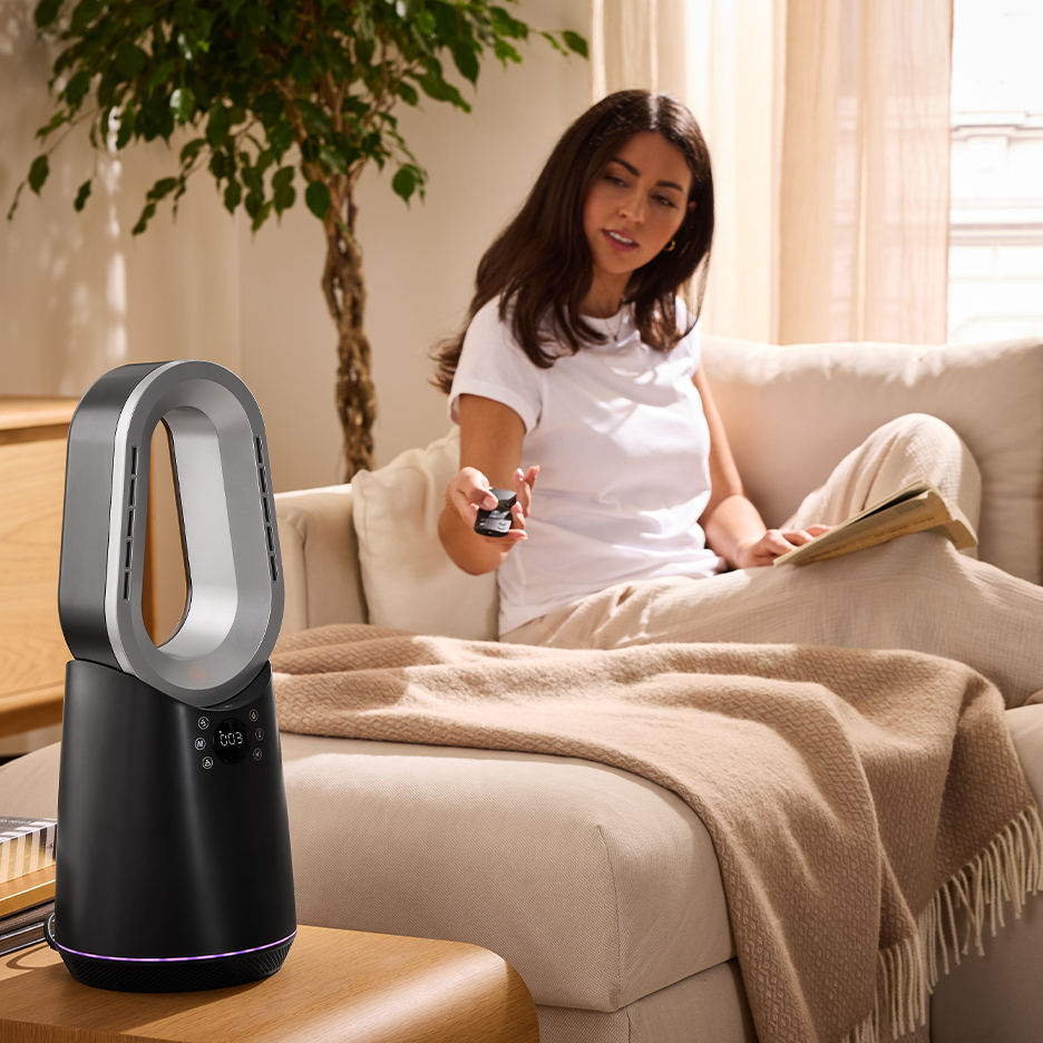 Una donna su un divano usa un telecomando per controllare un ventilatore senza pale nero e argento.