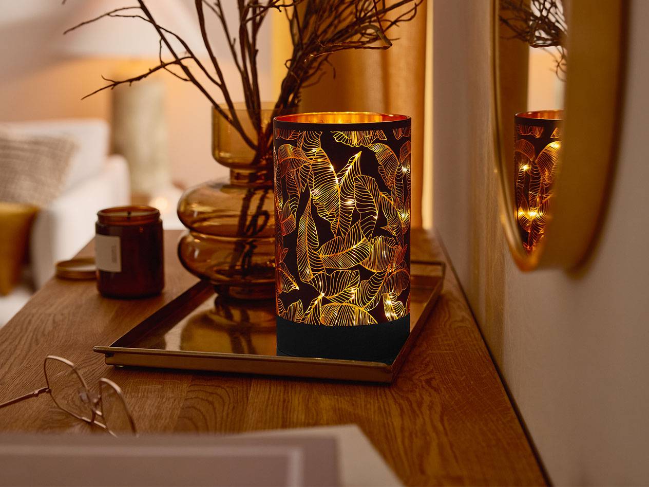Una lanterna LED decorativa con motivo a foglie, un vaso con rami e una candela su un tavolo di legno.
