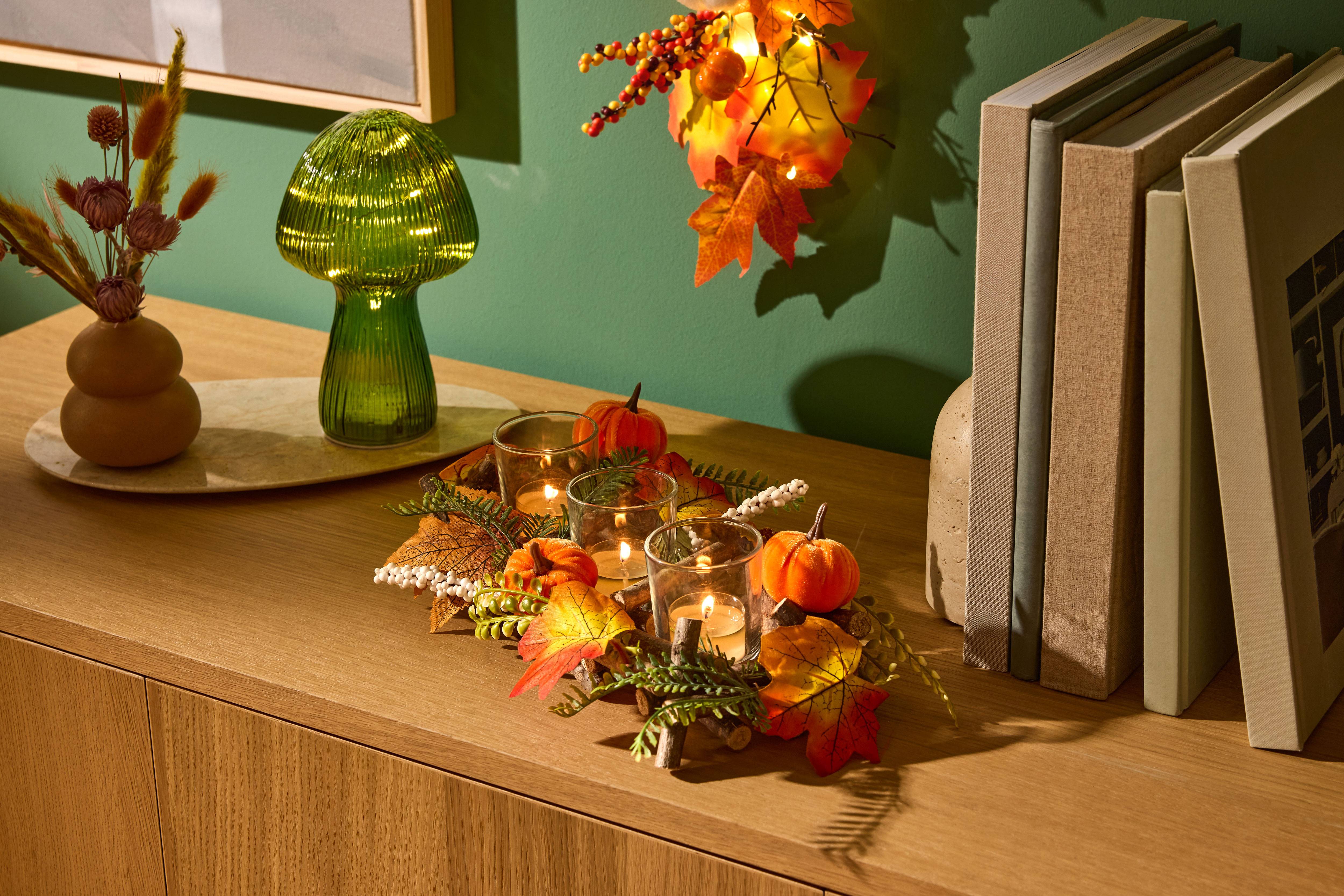 Decorazione autunnale da tavolo con zucche, candele e foglie, accanto a una lampada a fungo verde e libri.