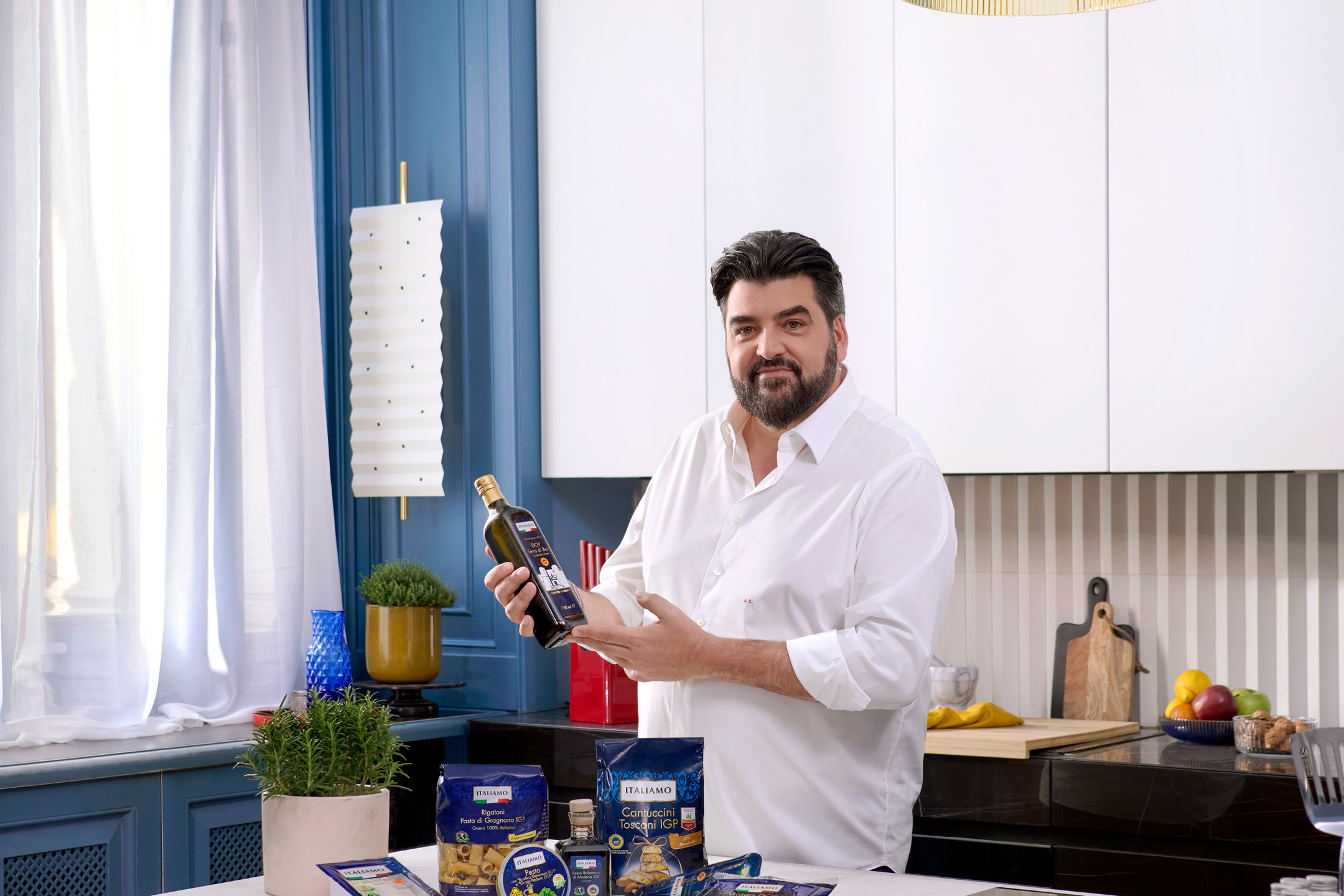 Uomo in cucina che mostra una bottiglia di olio d'oliva, con pasta, pesto e cantuccini sul tavolo.
