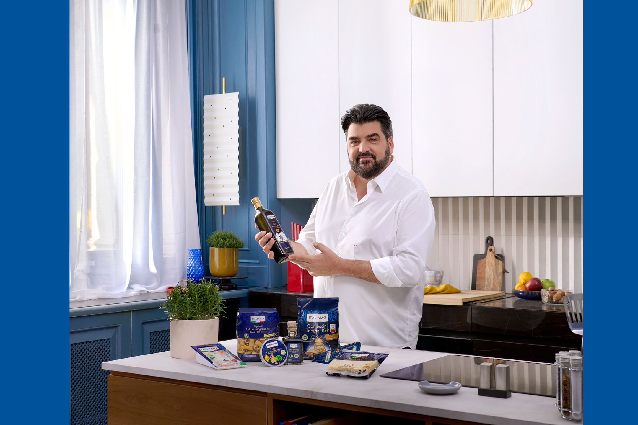 Uomo in cucina che mostra una bottiglia di olio d'oliva, con pasta, pesto e cantuccini sul bancone.