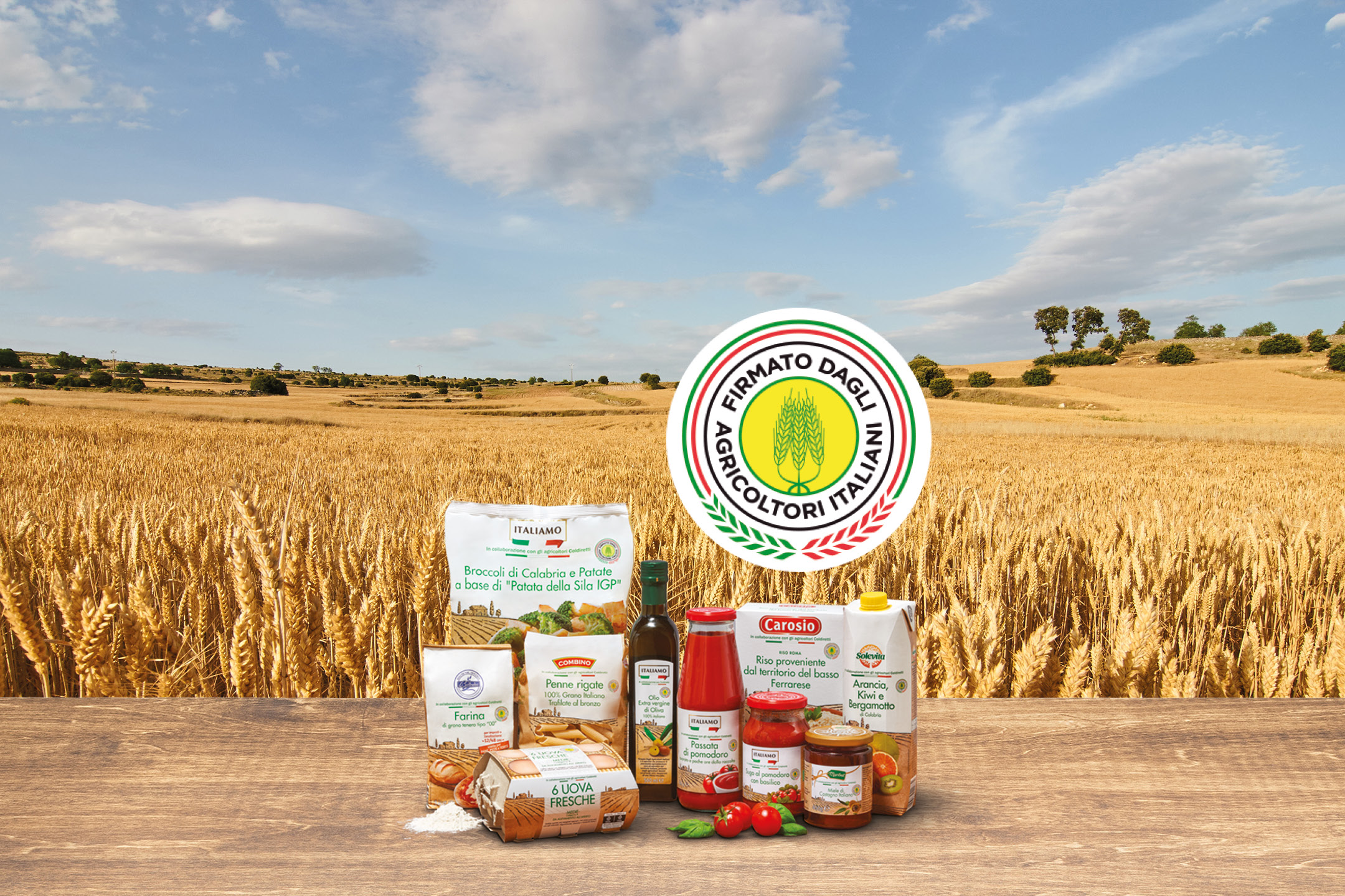 Prodotti alimentari italiani su un tavolo di legno con un campo di grano e cielo azzurro sullo sfondo.
