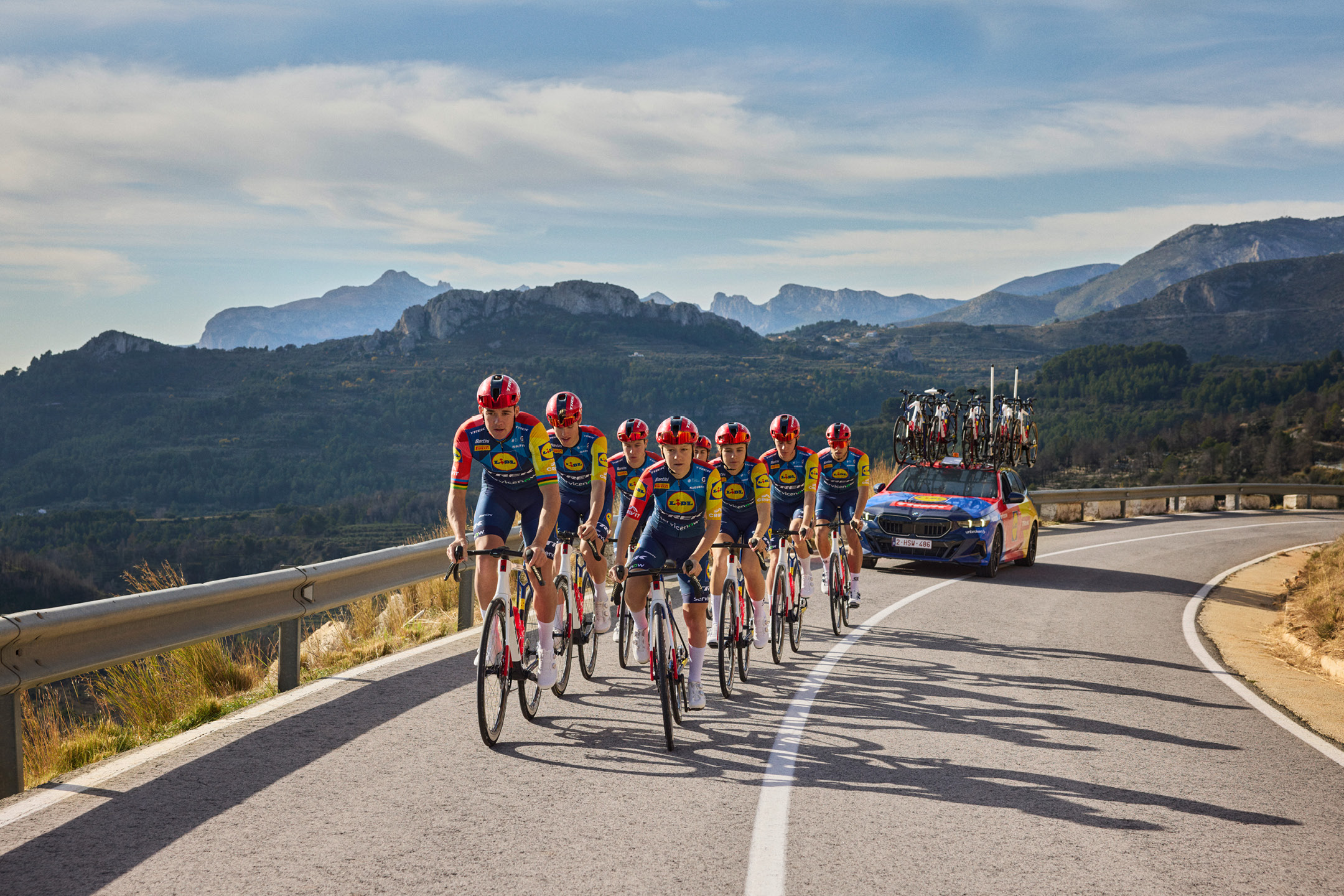 Ciclisti in maglia blu e gialla su strada di montagna, con auto di supporto e biciclette sul tetto.