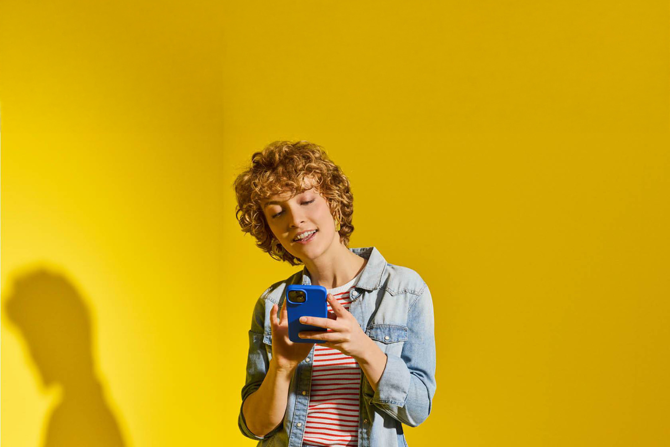 Donna sorridente con capelli ricci che tiene in mano uno smartphone blu su sfondo giallo.