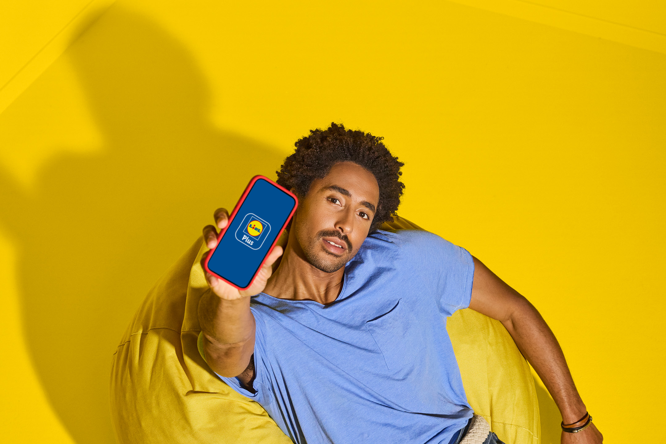 Uomo con maglietta blu che mostra un telefono con l'app Plus su sfondo giallo.