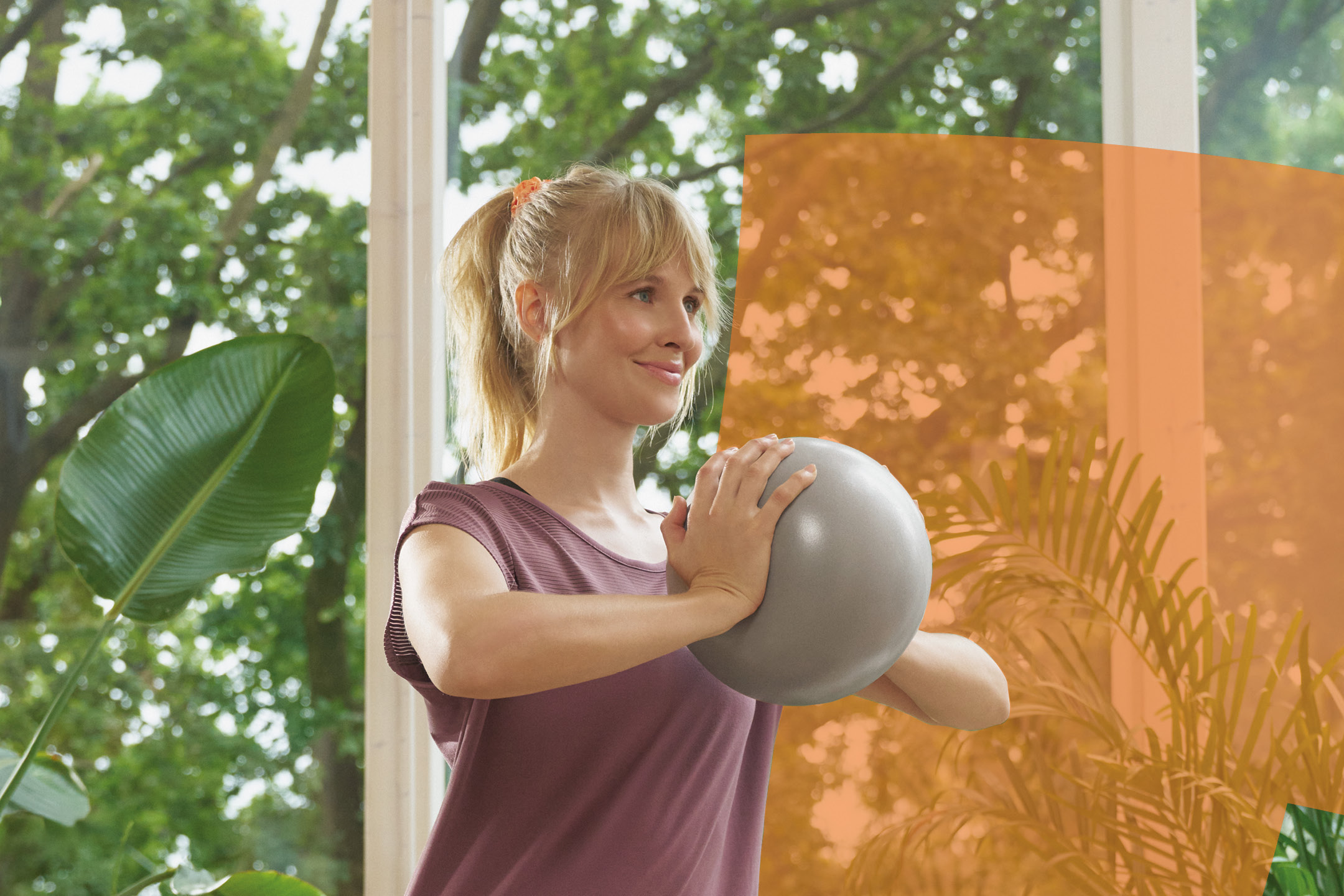 Donna che fa esercizio con una palla da pilates in un ambiente luminoso e verde.