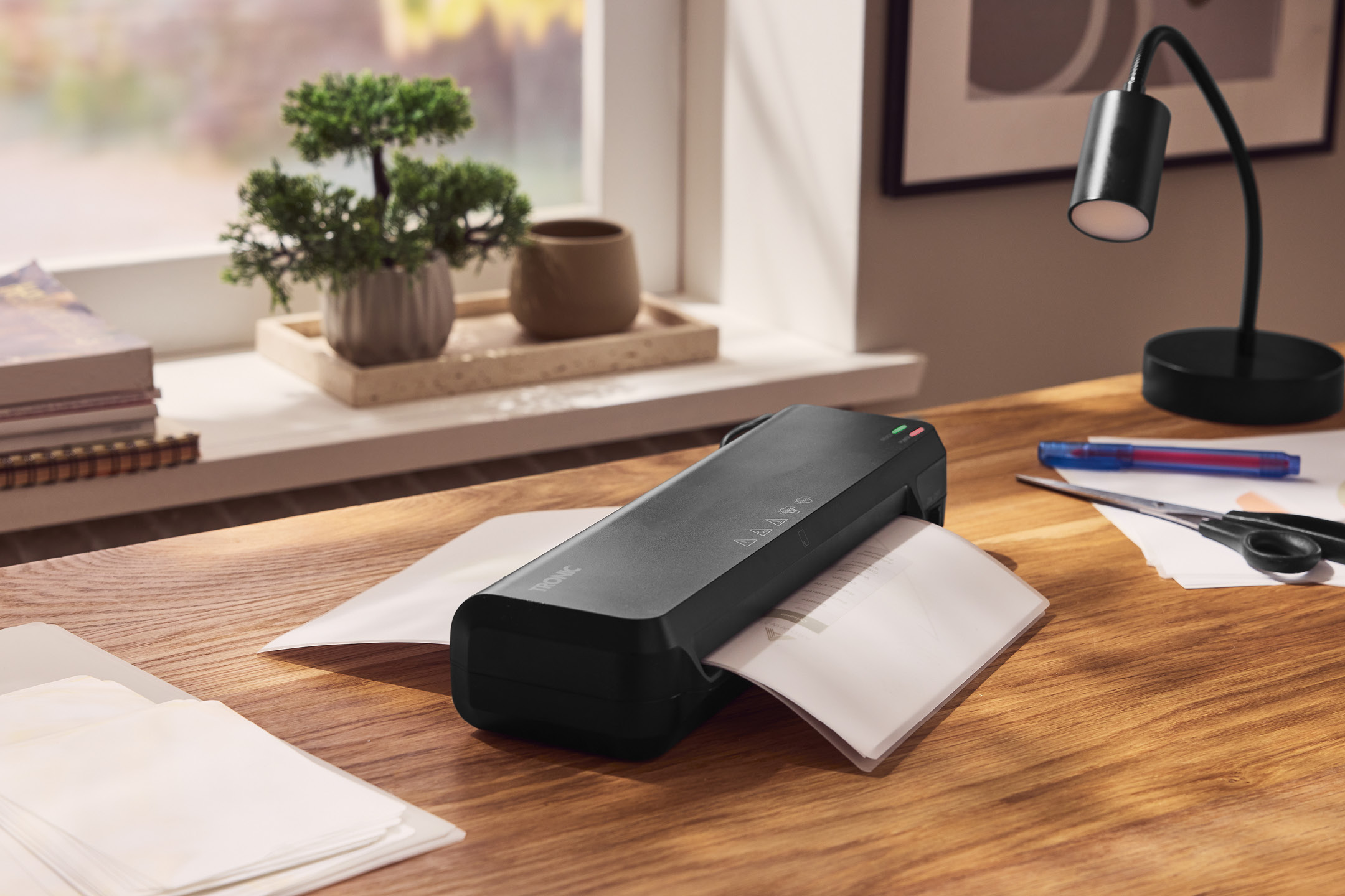 Plastificatore TRONIC nera su scrivania in legno con documenti, forbici e lampada da tavolo.
