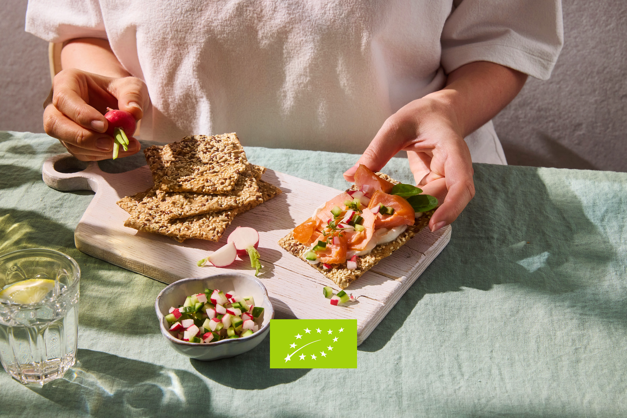Persona che prepara cracker con salmone, ravanelli e cetrioli su un tagliere di legno.