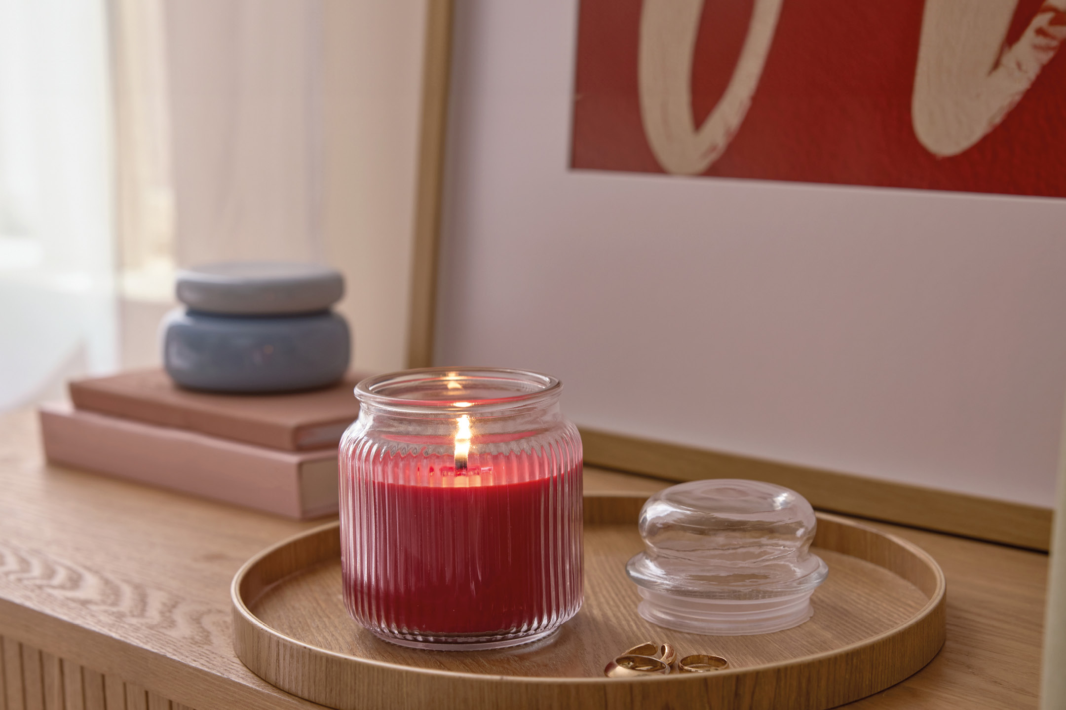 Candela rossa accesa in un barattolo di vetro scanalato su un vassoio di legno, con anelli e libri sullo sfondo.