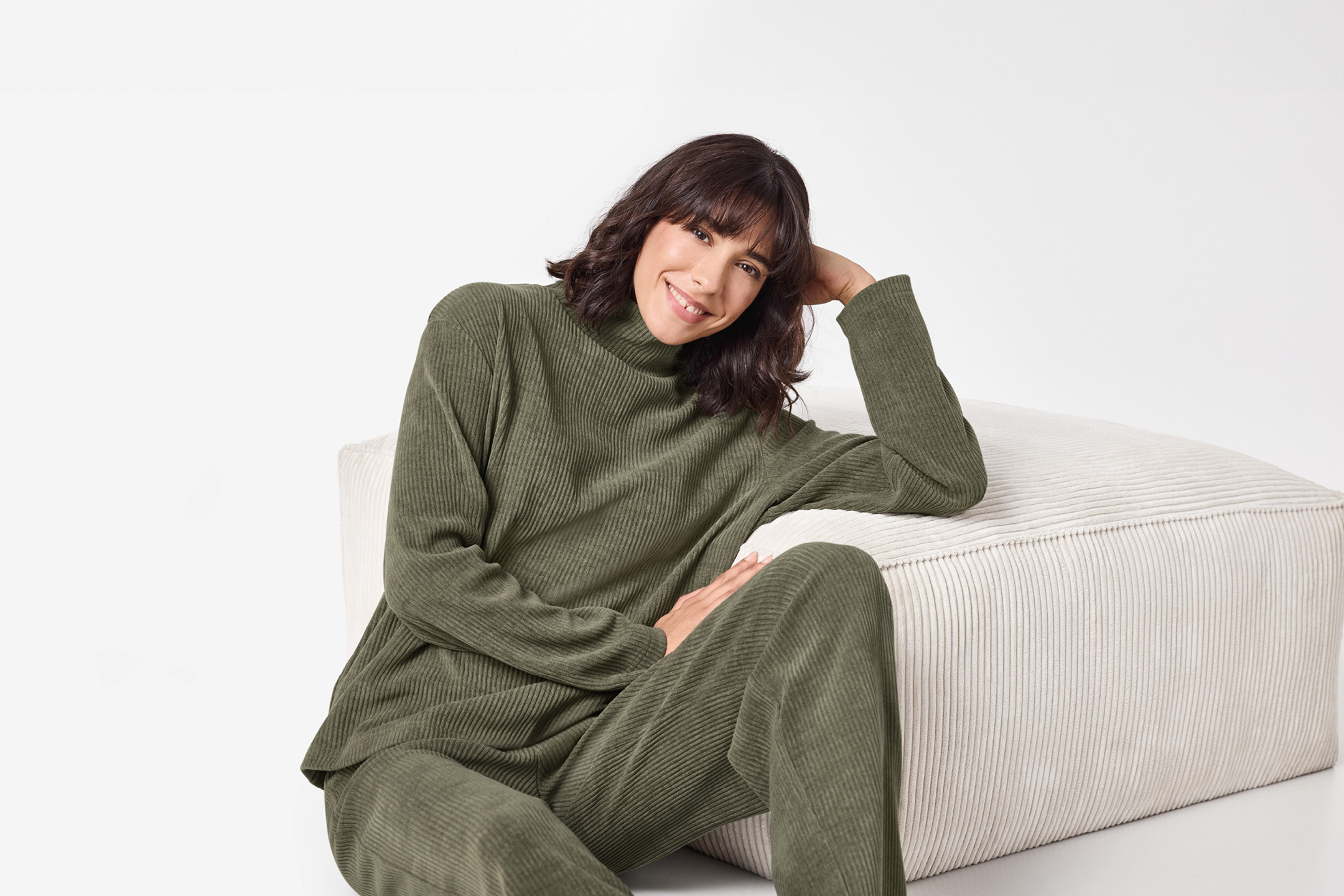Donna sorridente in completo loungewear a coste verde oliva, seduta accanto a un pouf.