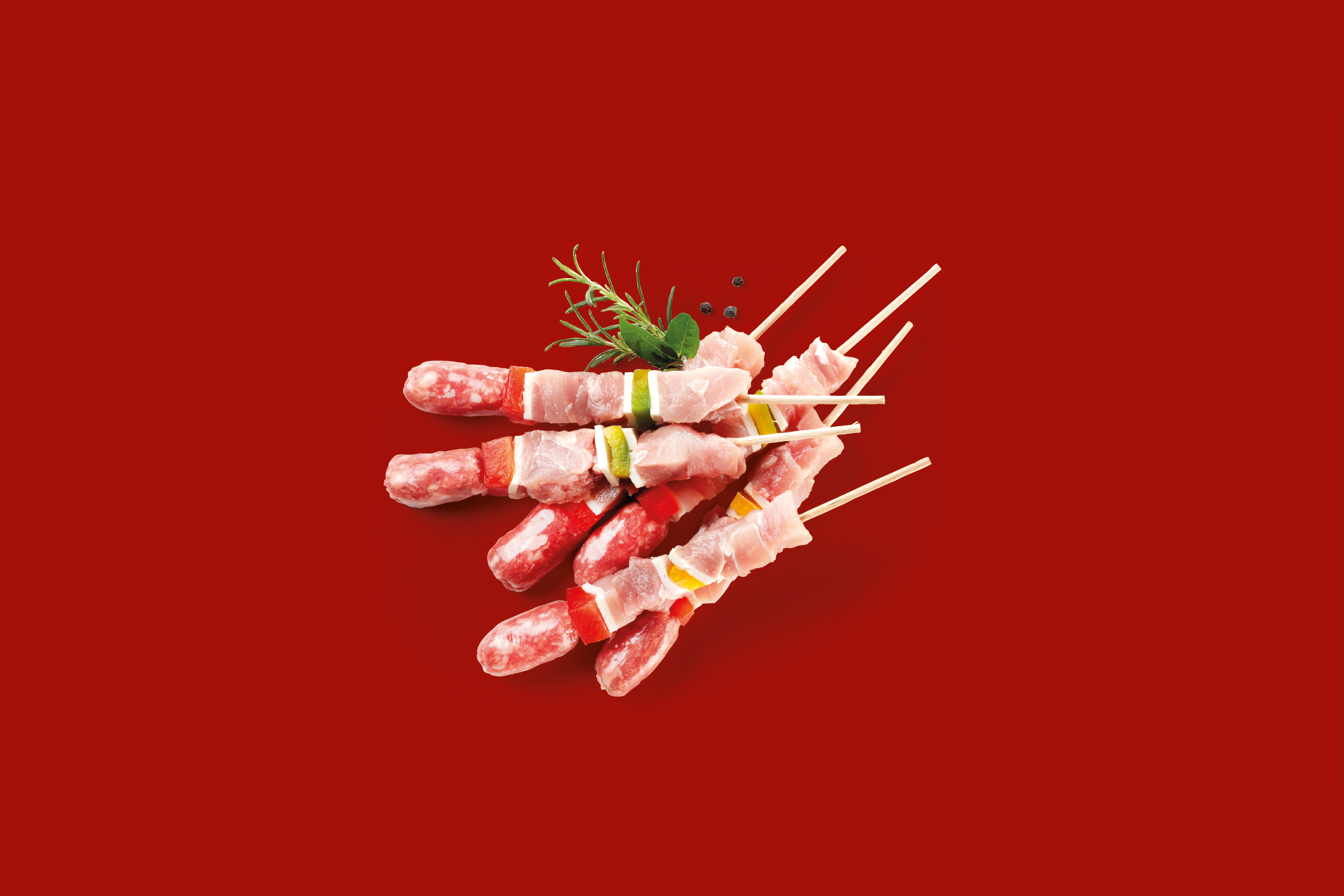 Spiedini di carne e salsiccia con verdure e rosmarino su sfondo rosso.