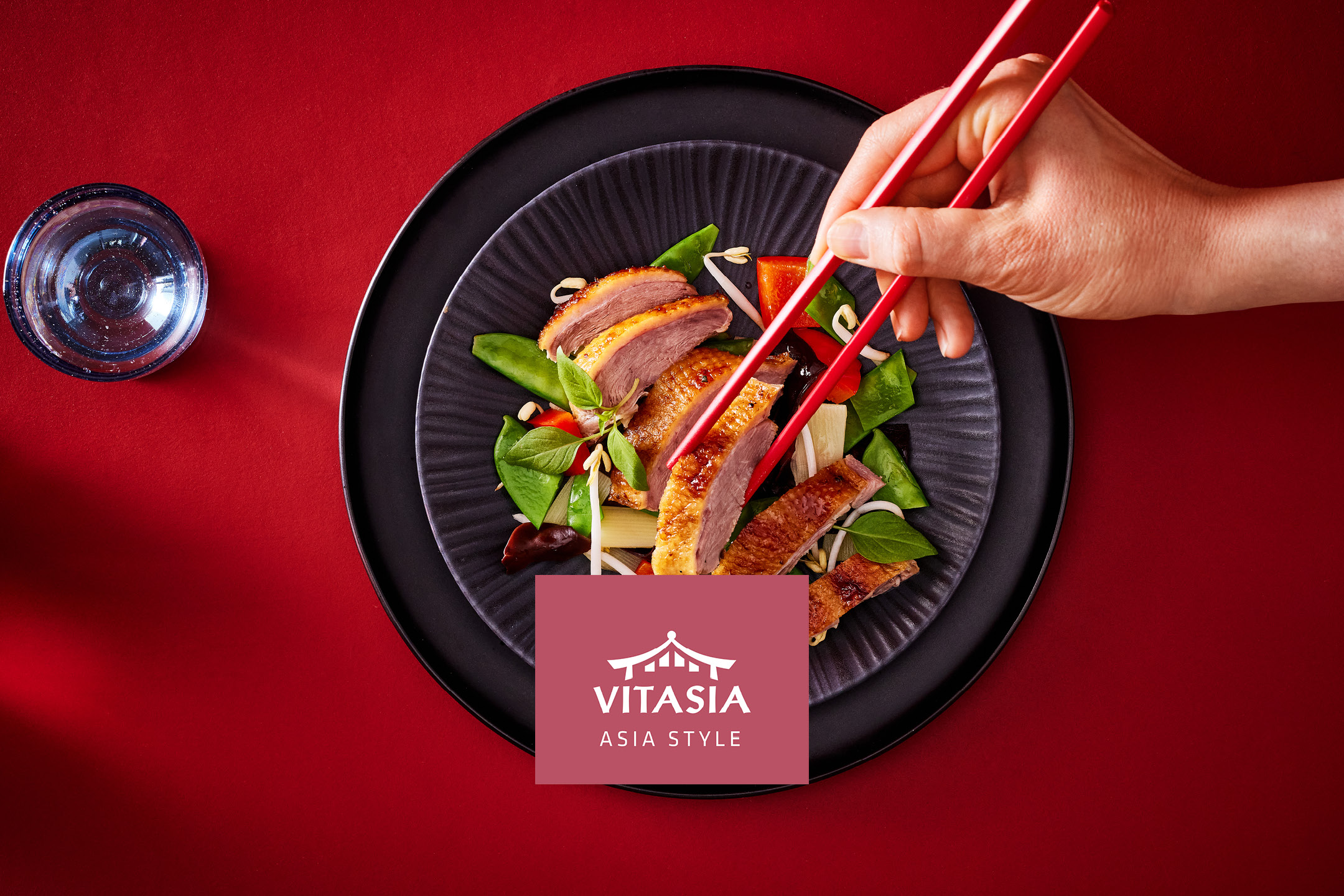 Anatra asiatica con verdure e bacchette su un piatto nero, con un bicchiere d'acqua.