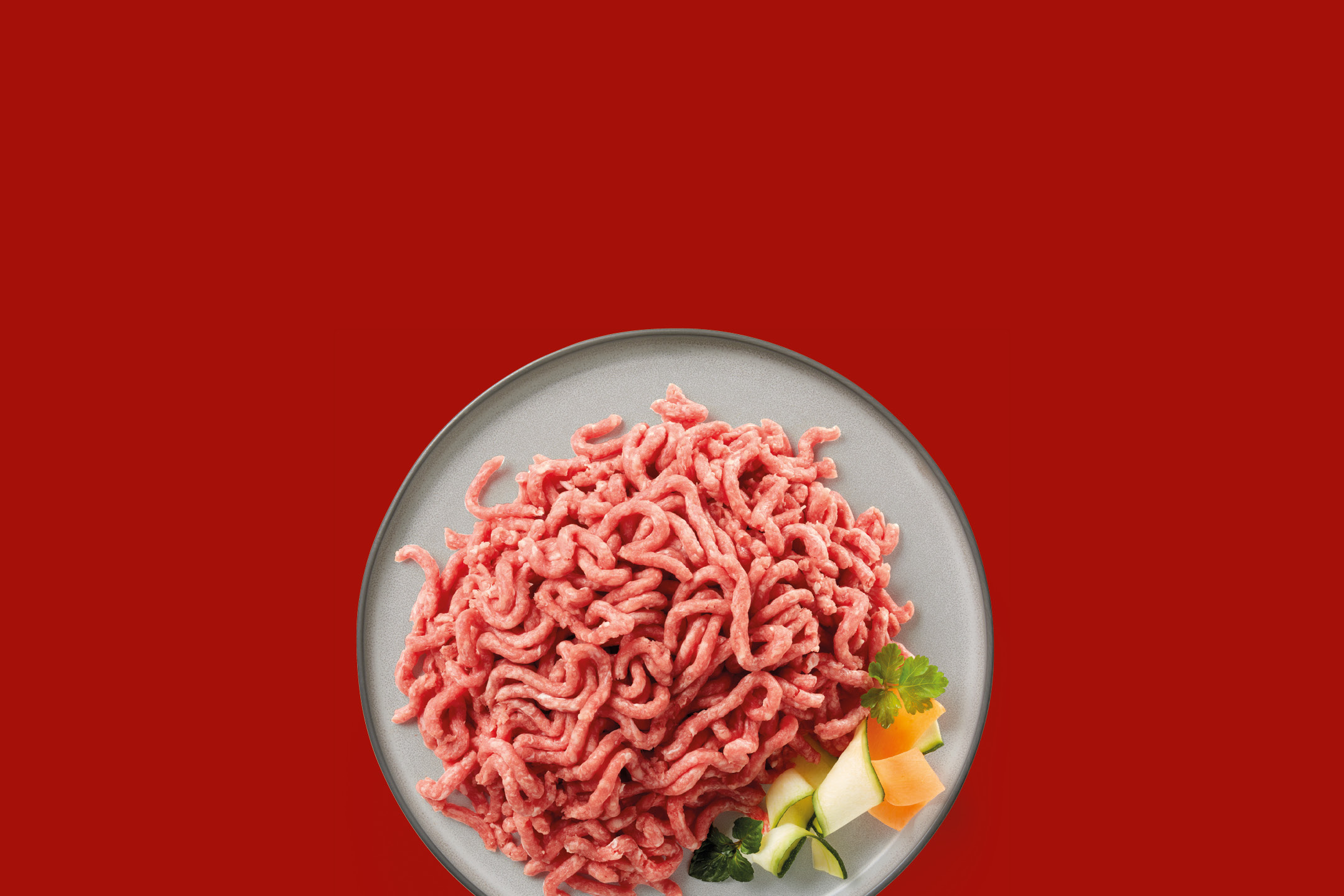 Carne macinata cruda su un piatto grigio con contorno di verdure su sfondo rosso.