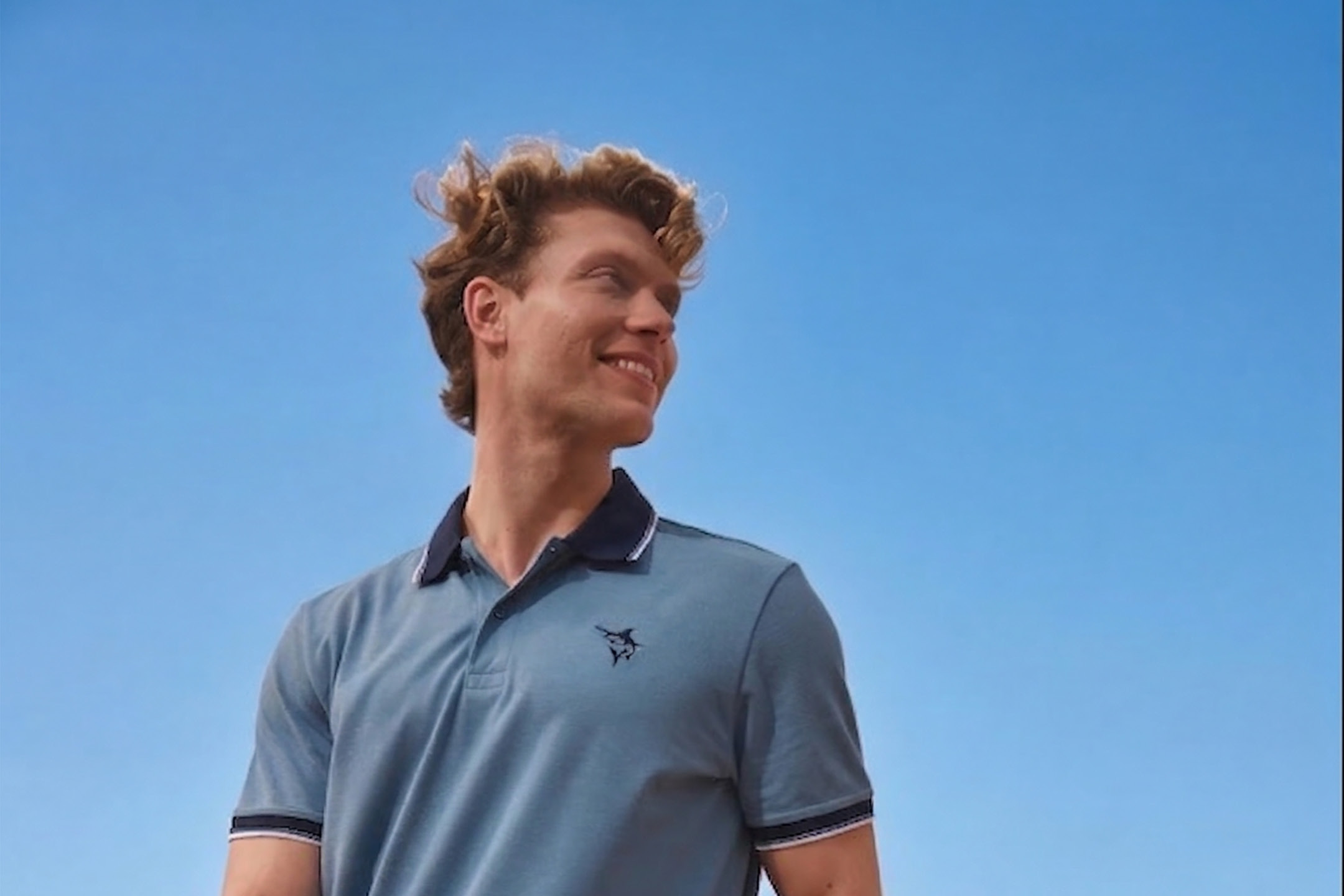 Uomo sorridente con polo blu e colletto a contrasto, su sfondo di cielo azzurro.