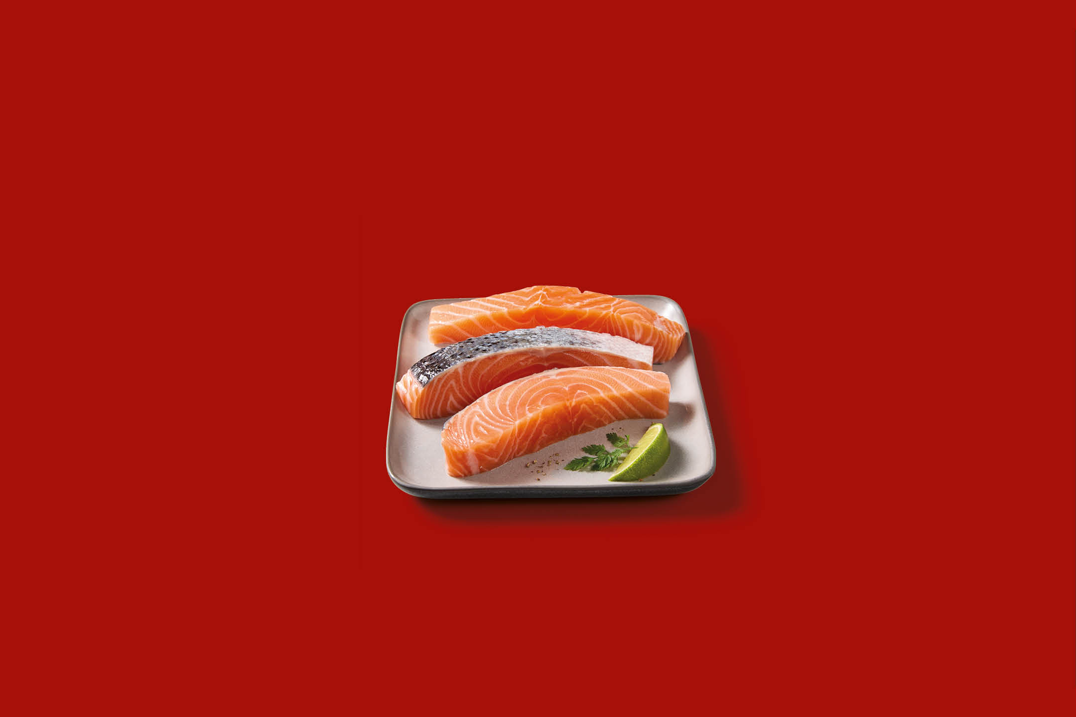 Filetti di salmone fresco su un piatto con lime e prezzemolo, su sfondo rosso.
