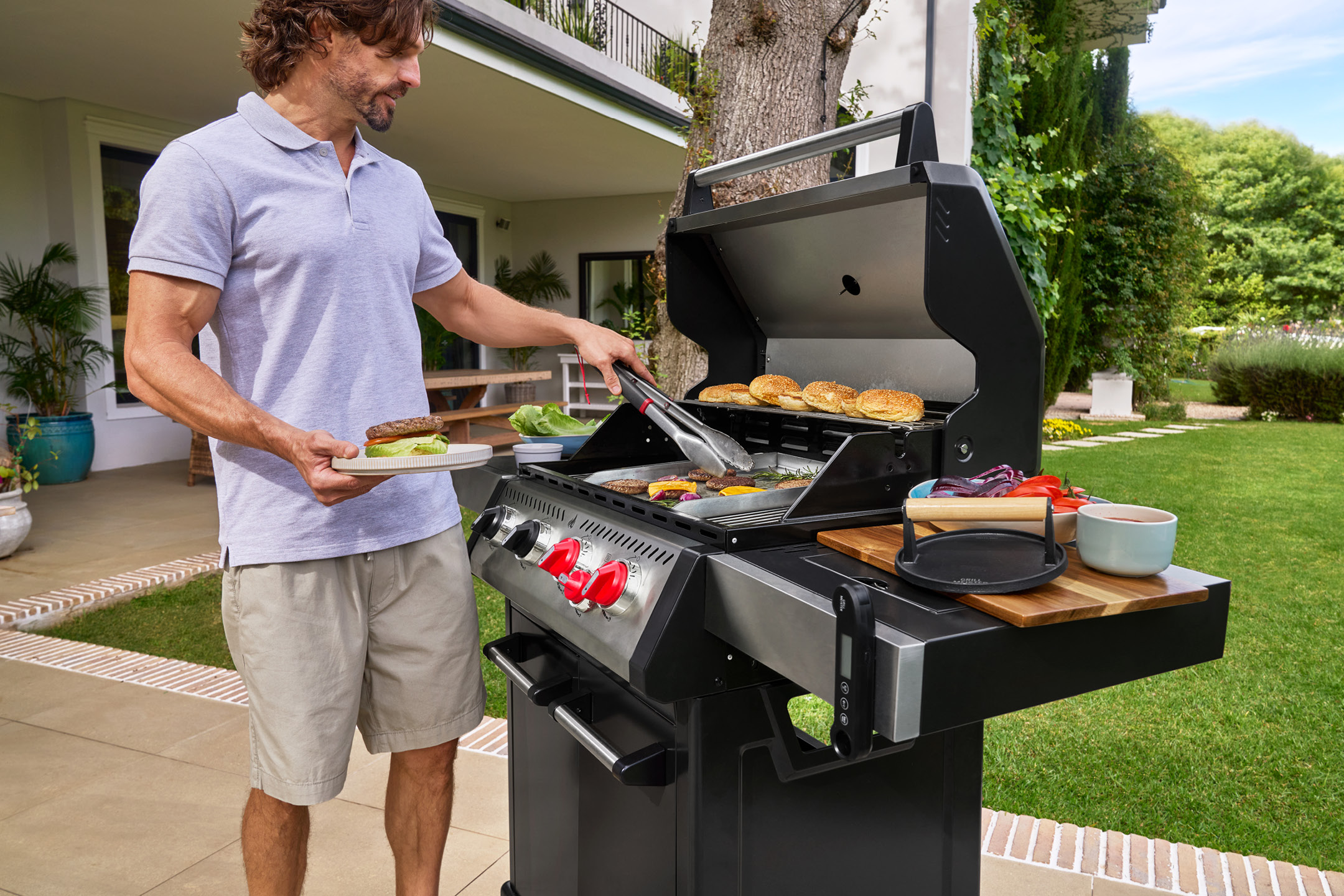 Uomo che griglia hamburger e panini su un barbecue a gas in un giardino.