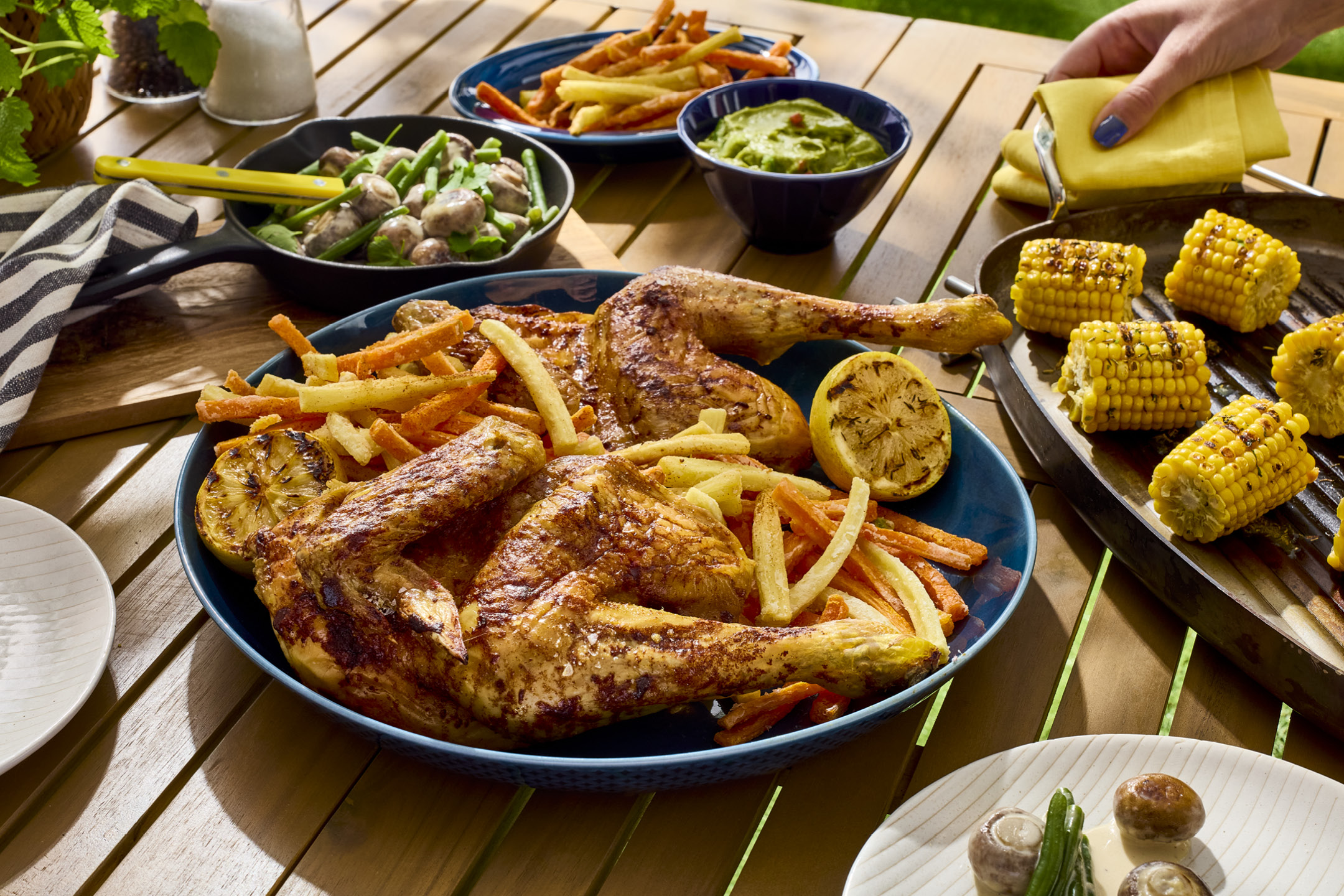 Pollo arrosto con patatine fritte, pannocchie di mais grigliate e contorni su un tavolo di legno.