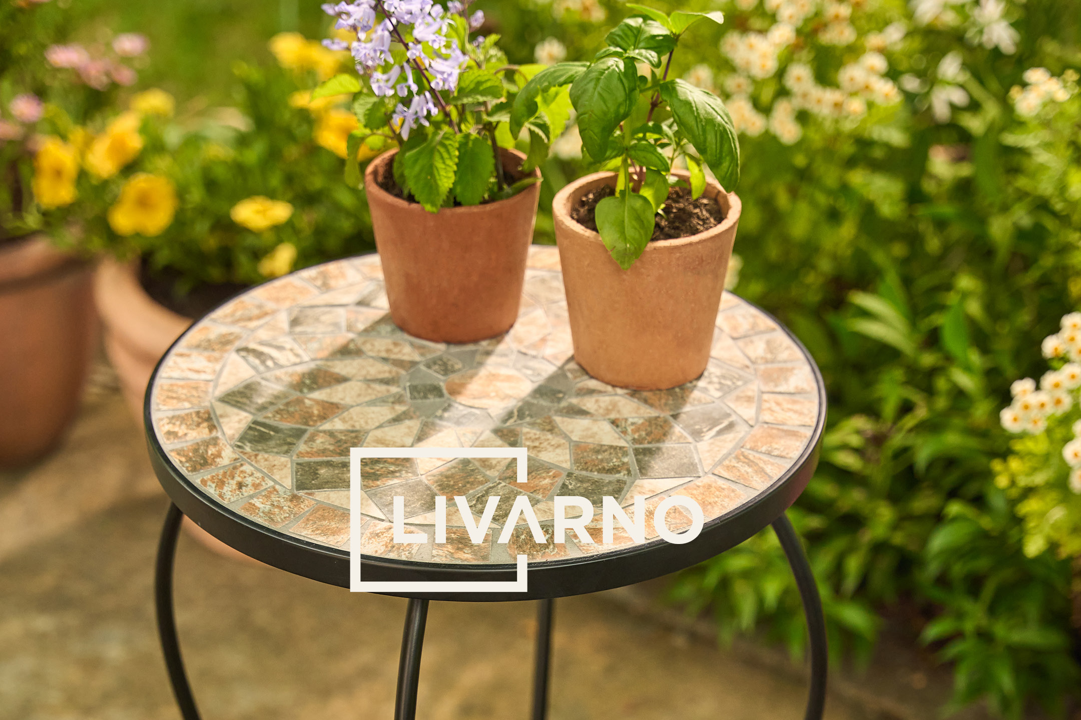Tavolino da giardino con mosaico e due piante in vaso, sullo sfondo un giardino fiorito.