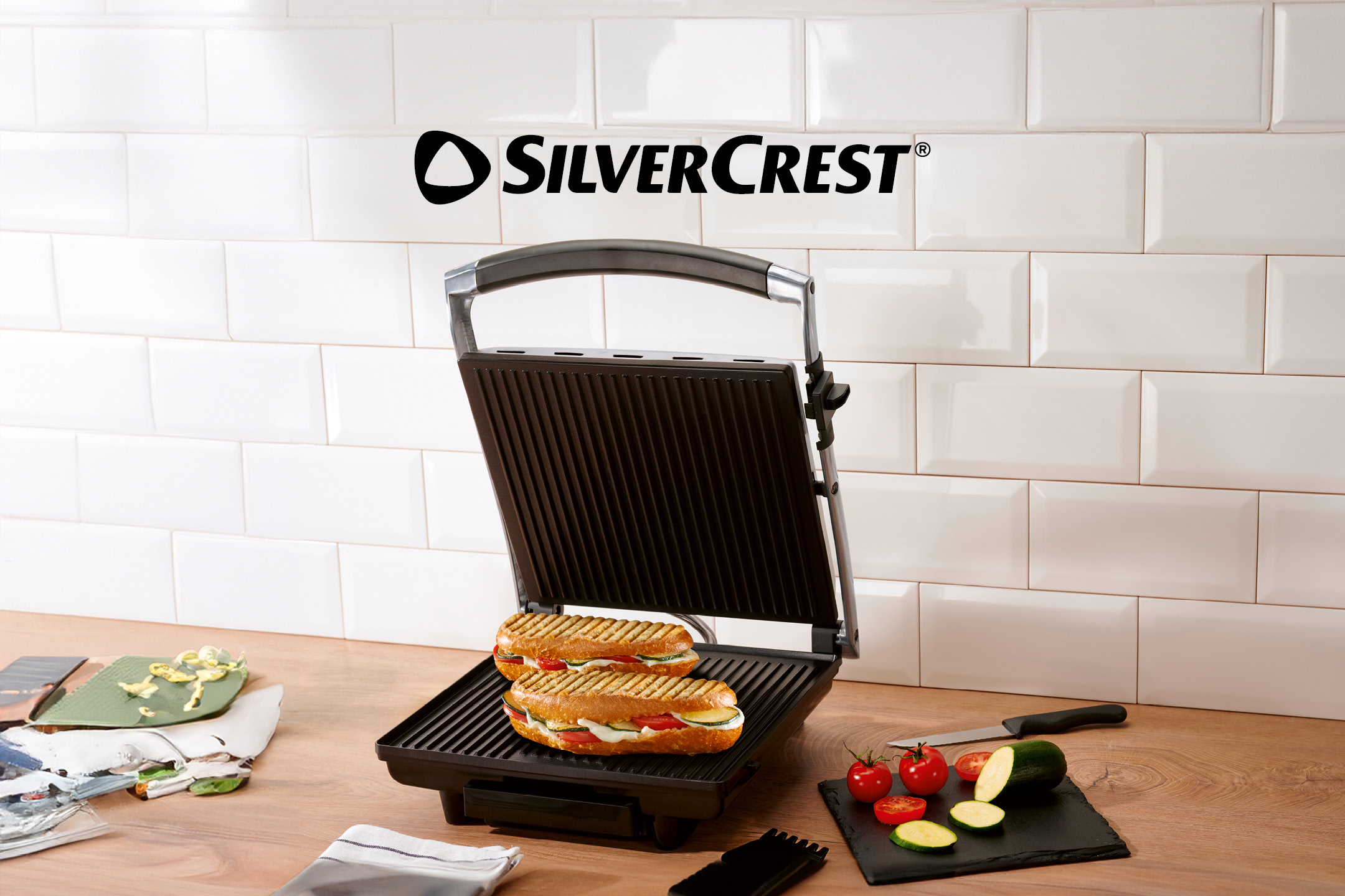 Piastra per panini aperta con due panini farciti, verdure fresche e utensili da cucina su un tavolo di legno.