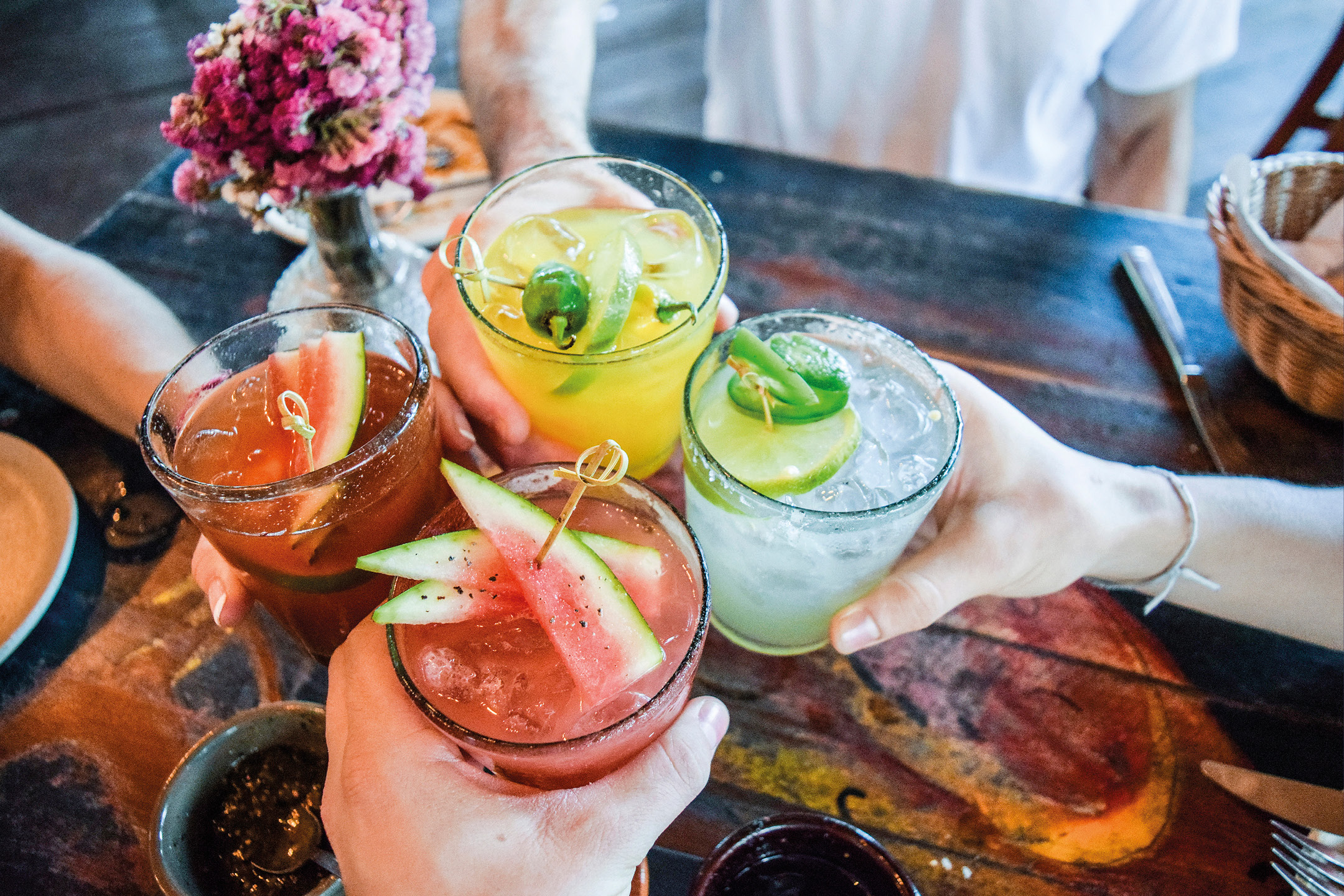 Quattro persone brindano con cocktail colorati guarniti con frutta e peperoncini.