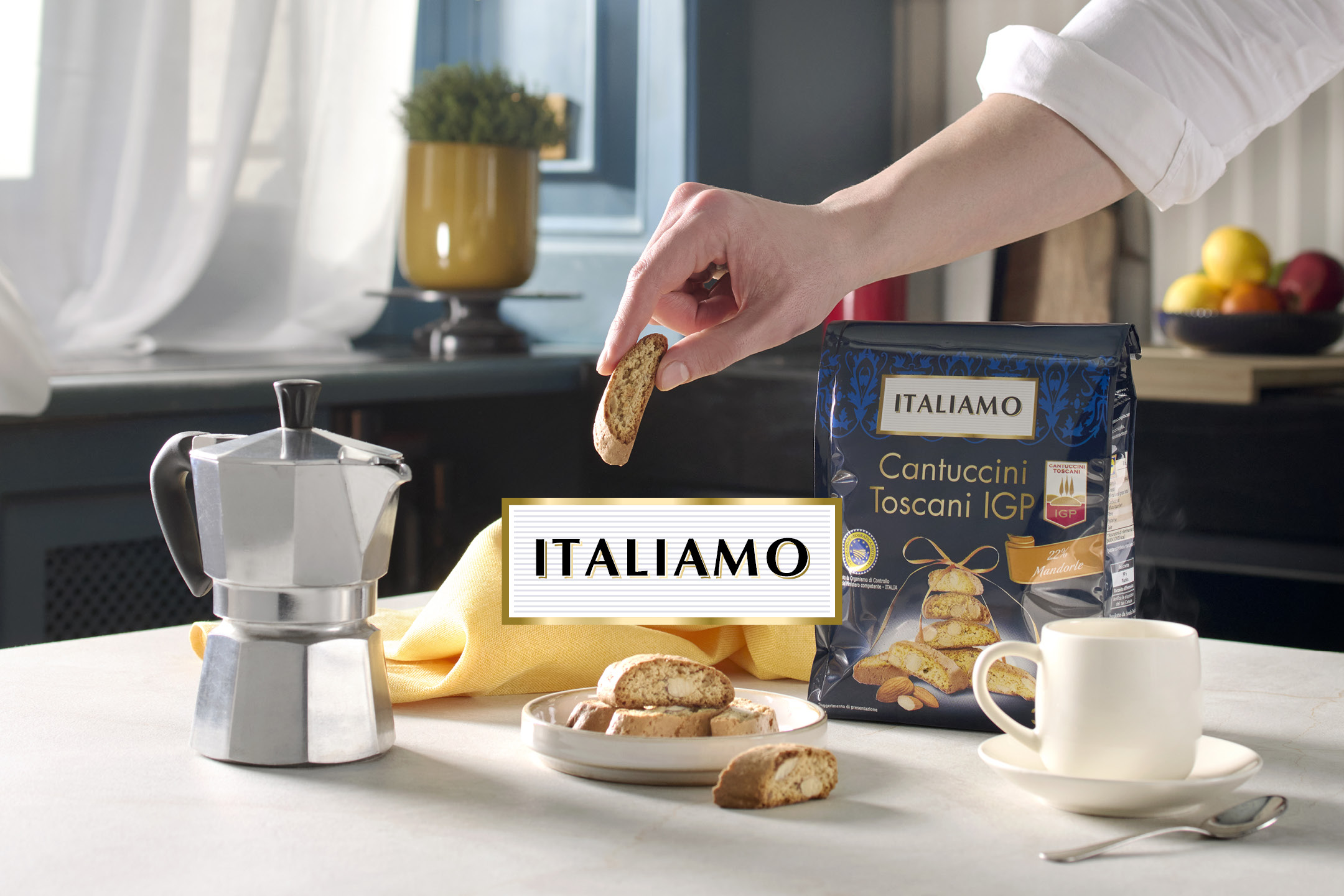 Mano che prende un cantuccino da un piatto, con una moka e una confezione di biscotti alle mandorle.