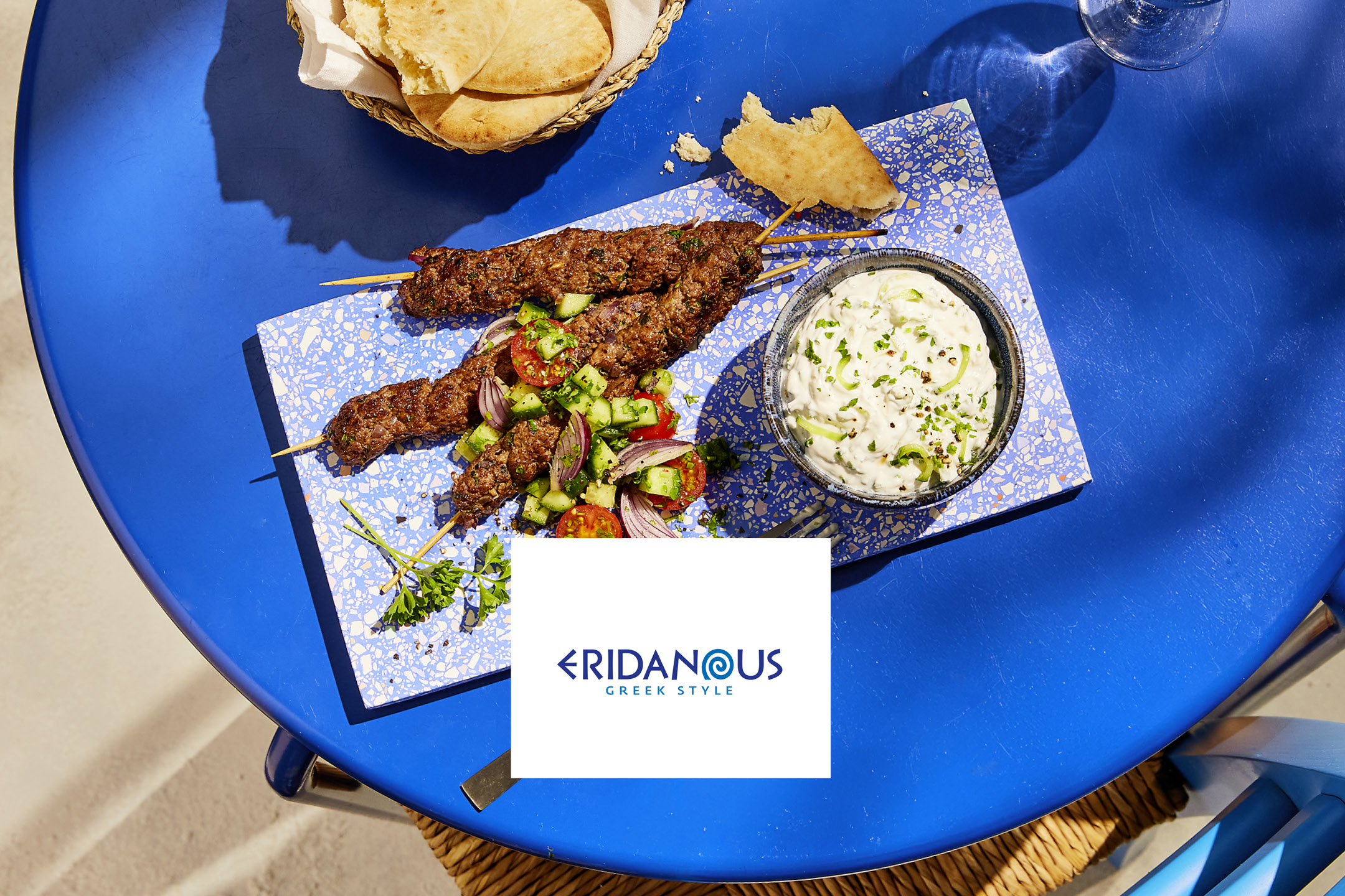 Spiedini di carne macinata con insalata di pomodori e cetrioli, tzatziki e pane pita su un tavolo blu.