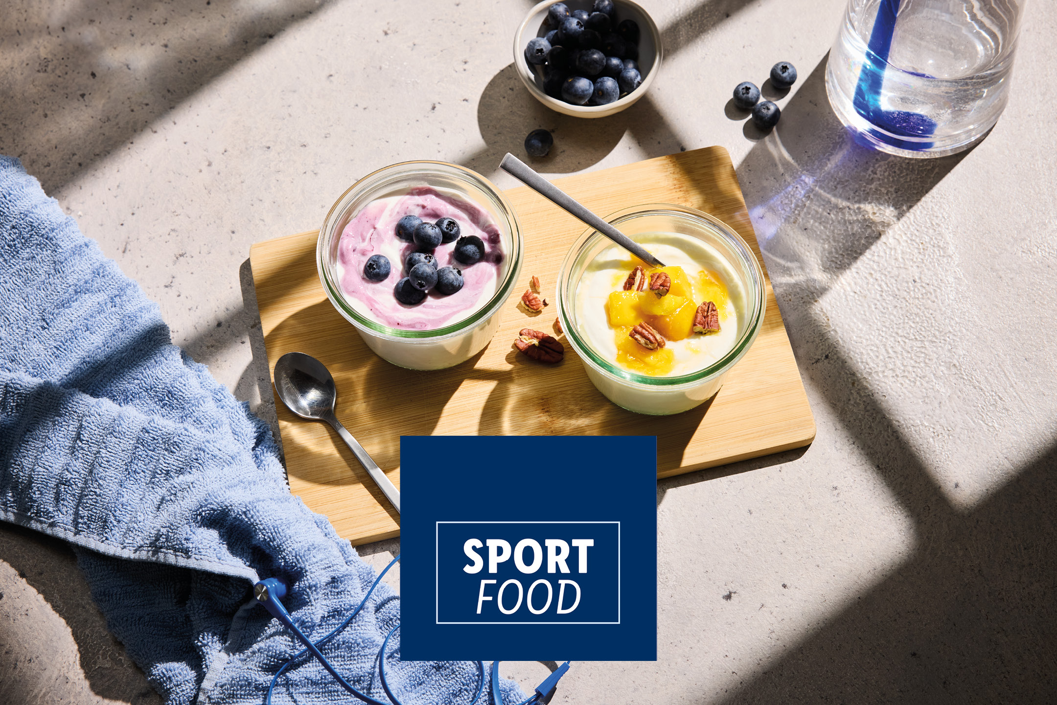 Due vasetti di yogurt con frutta e noci su un tagliere, con asciugamano e cuffie. Testo: SPORT FOOD.