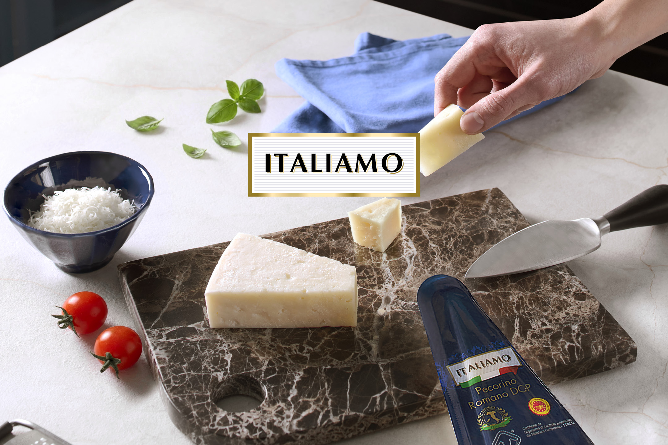 Formaggio grattugiato e a pezzi su tagliere di marmo, con pomodorini e coltello, con scritta 'ITALIAMO'.