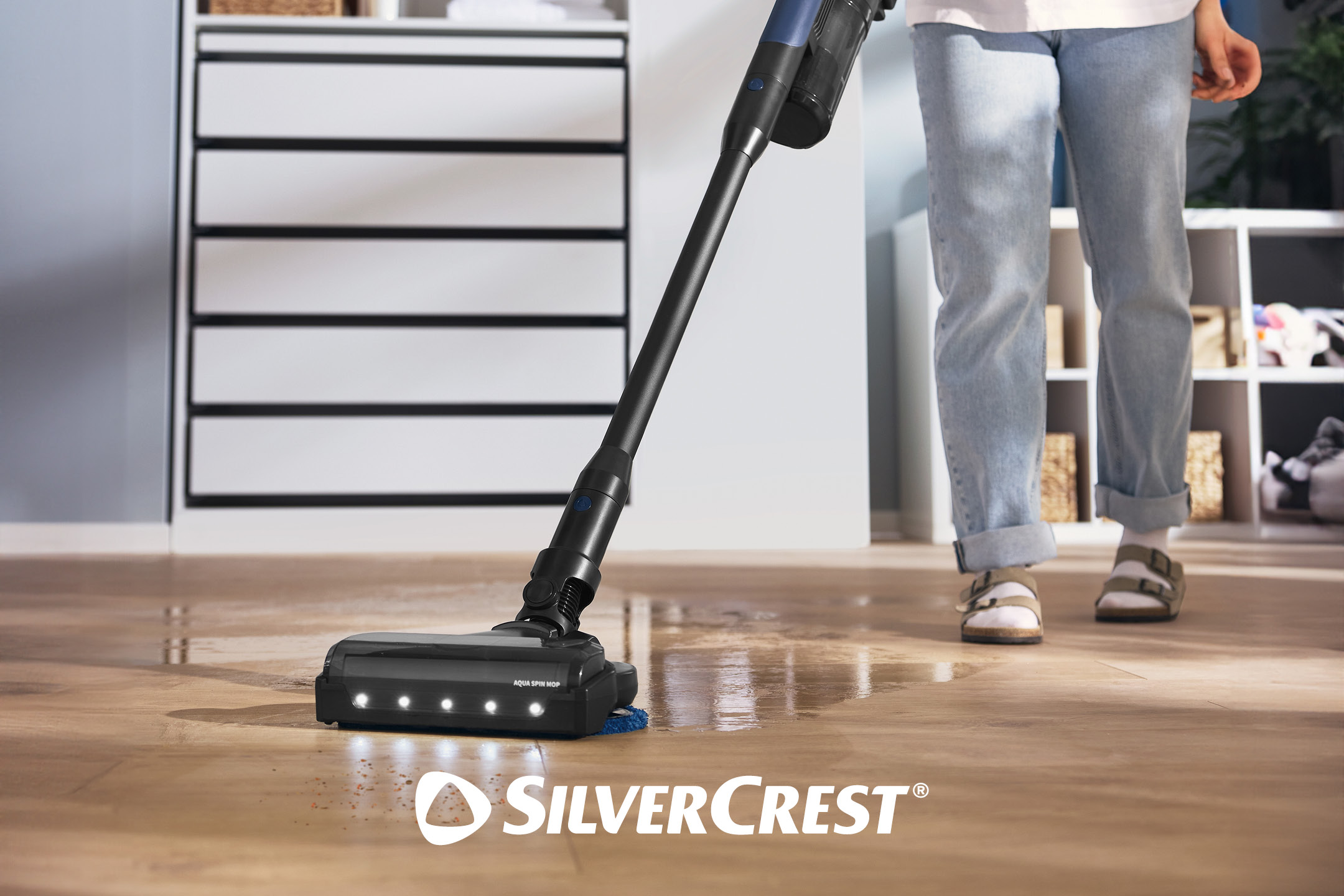 Mocio lavapavimenti elettrico che pulisce un pavimento in legno con una persona in piedi sullo sfondo.