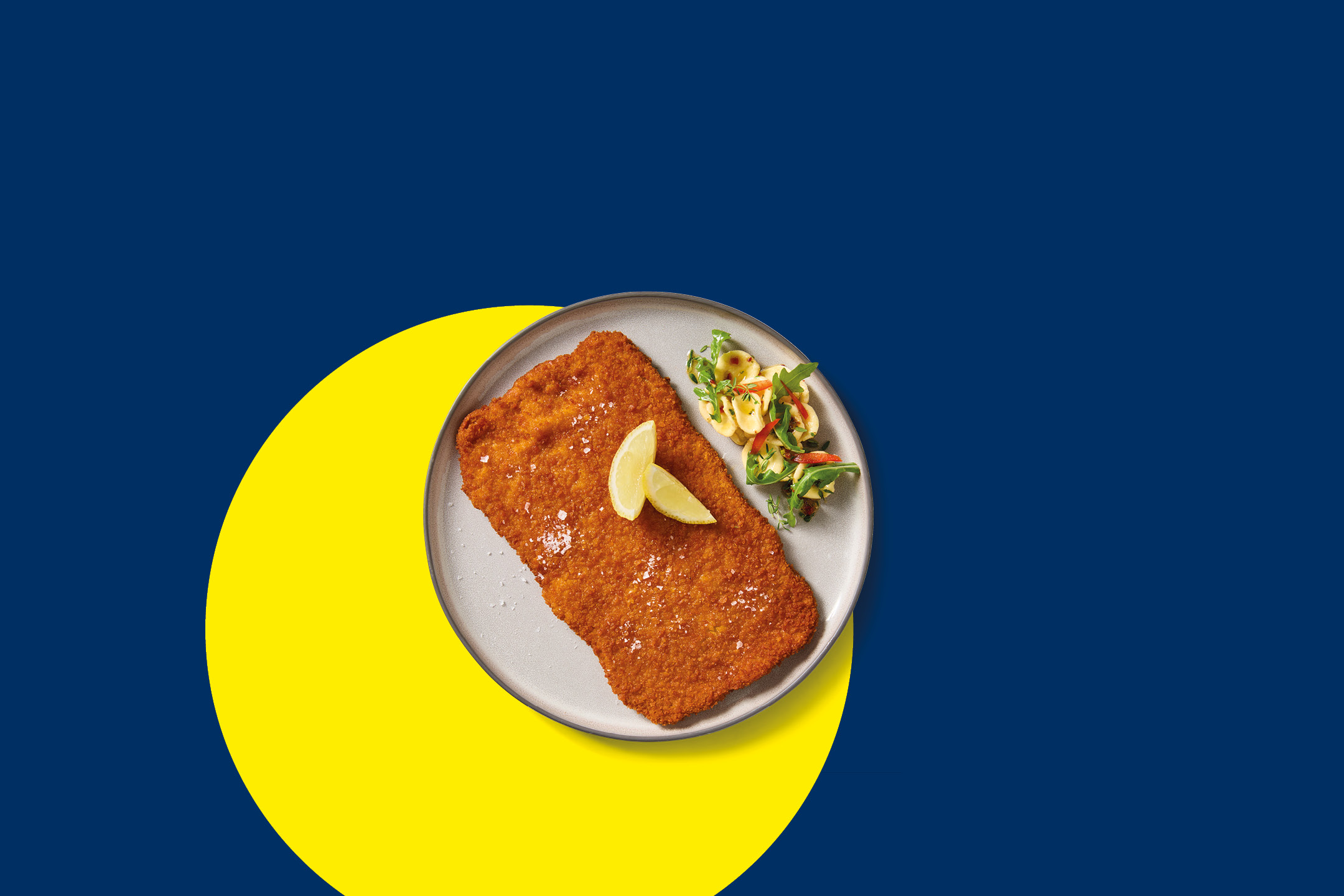 Cotoletta impanata con spicchi di limone e insalata su un piatto, sfondo blu e giallo.