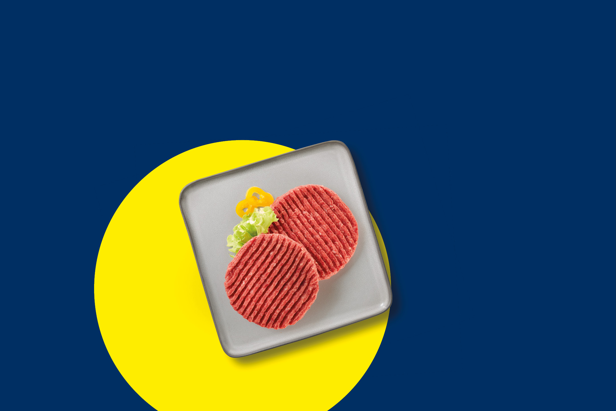 Due hamburger crudi su un piatto grigio con lattuga e peperoni gialli, su sfondo blu e giallo.
