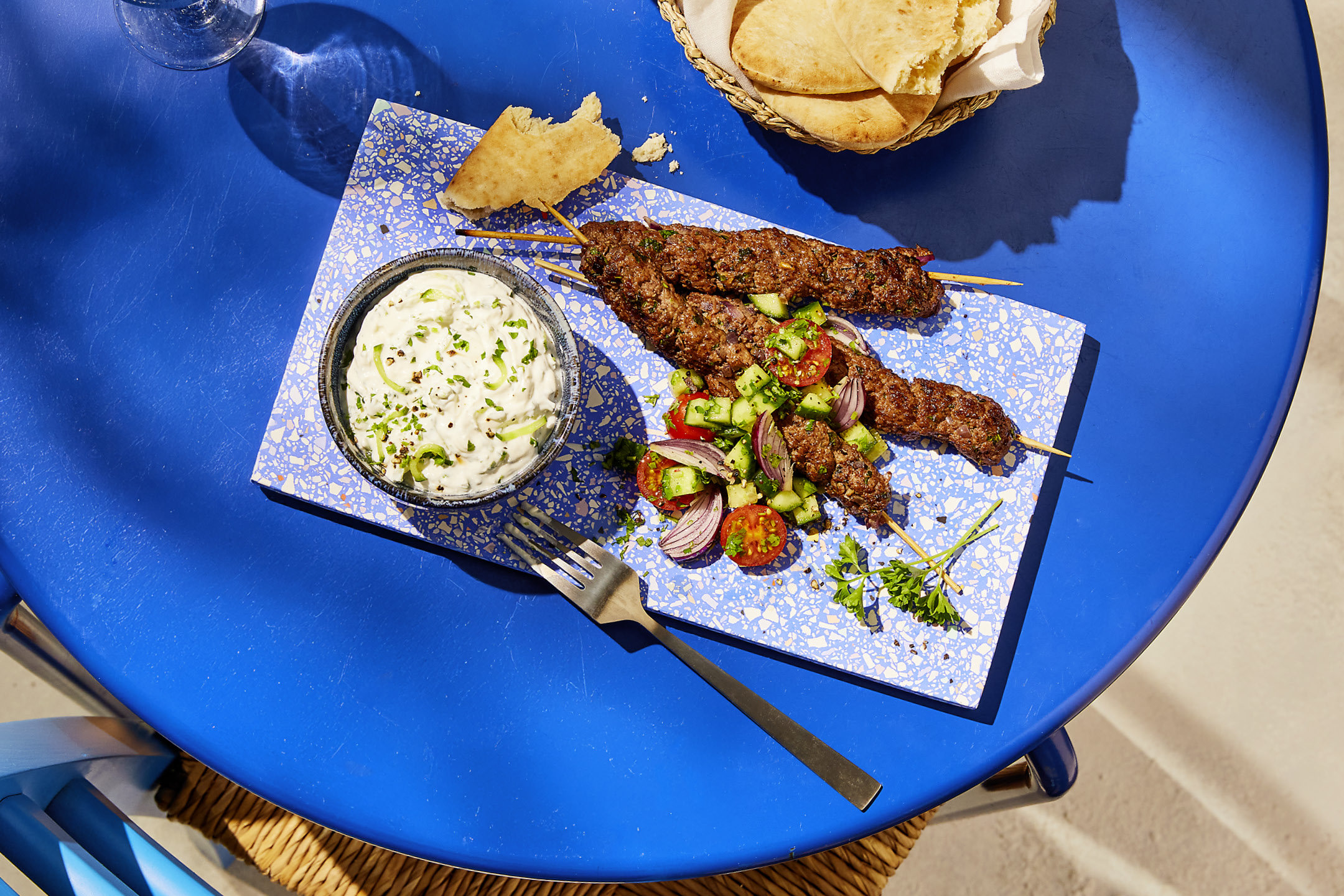 Spiedini di carne con salsa tzatziki, insalata di pomodori e cetrioli, e pane pita su un tavolo blu.
