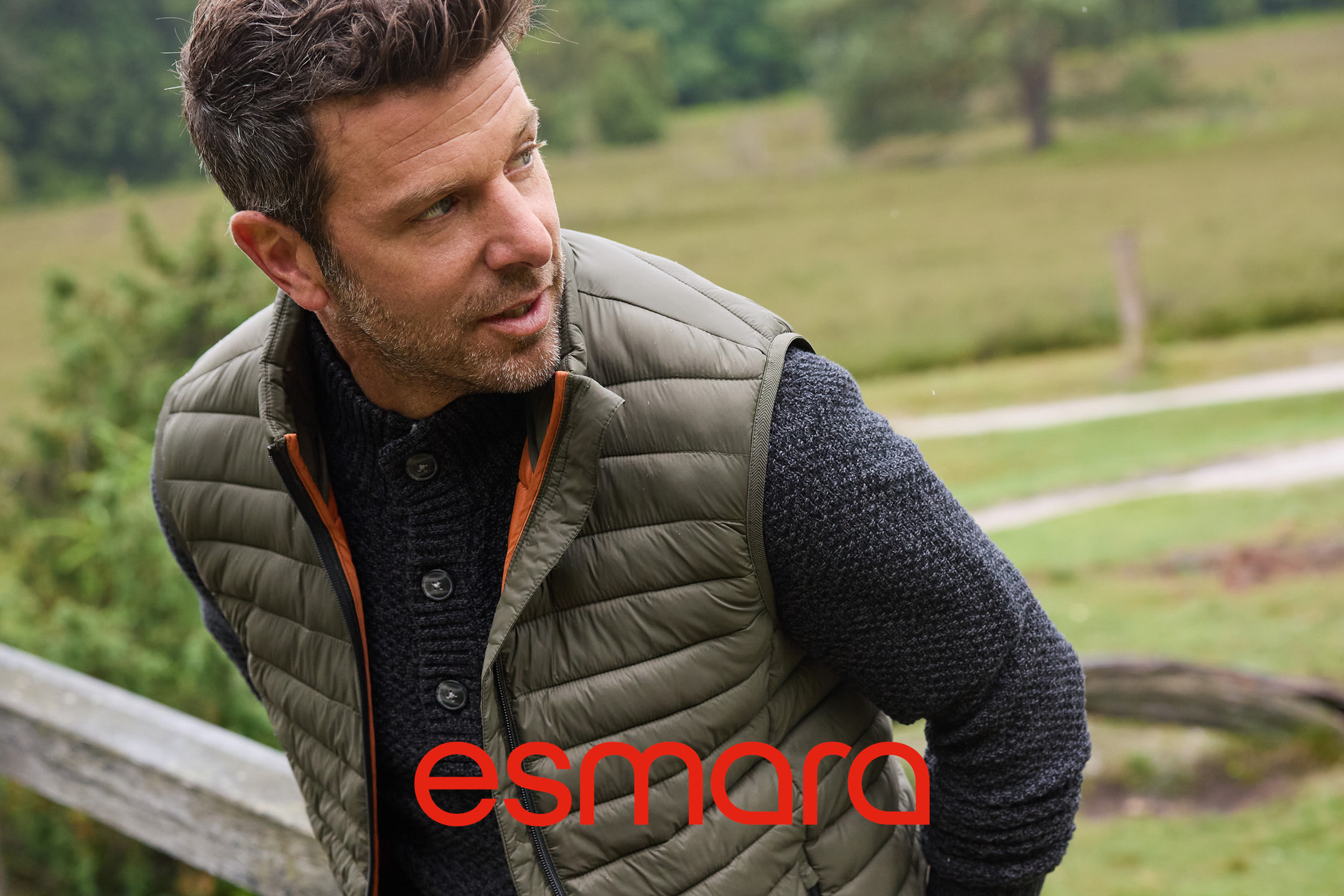 Uomo con gilet trapuntato verde oliva e maglione lavorato a maglia grigio scuro all'aperto.
